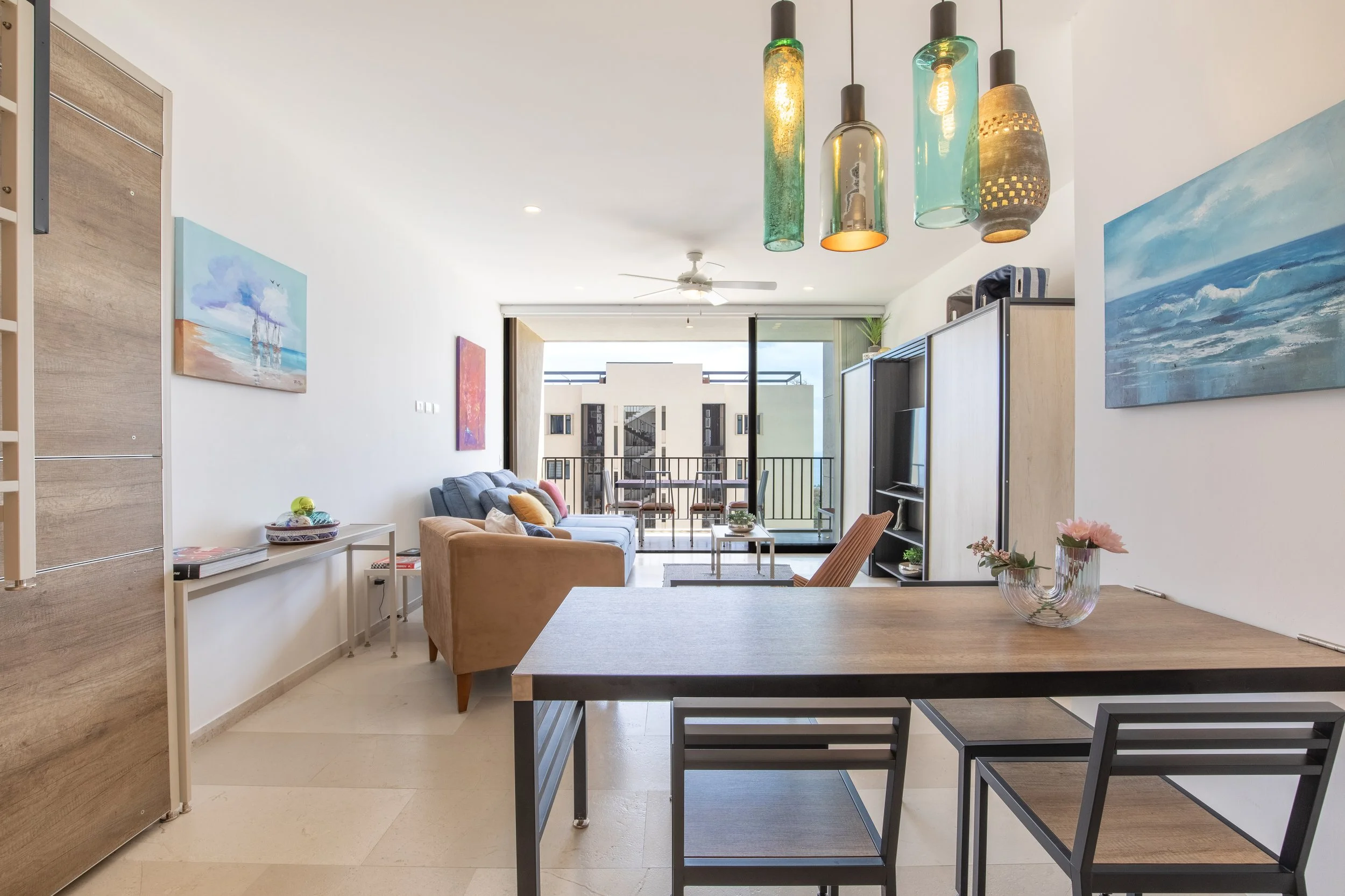 Modern living room with artwork on walls, a banquette with pillows, a glass coffee table, and hanging pendant lights over dining area, with balcony doors leading outside.