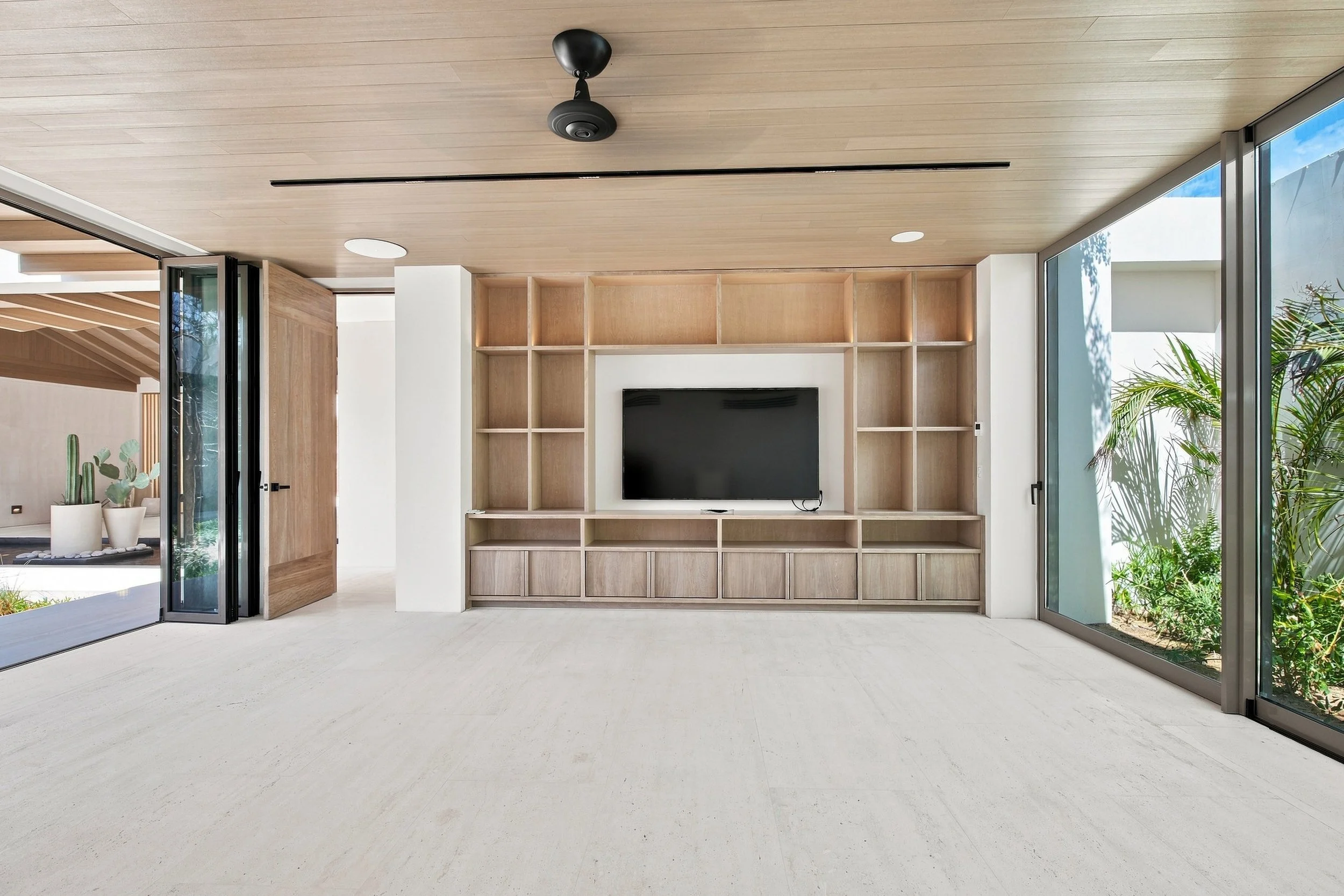 Modern living room with built-in wooden wall unit, flat-screen TV, large glass sliding doors, and potted plants outside.