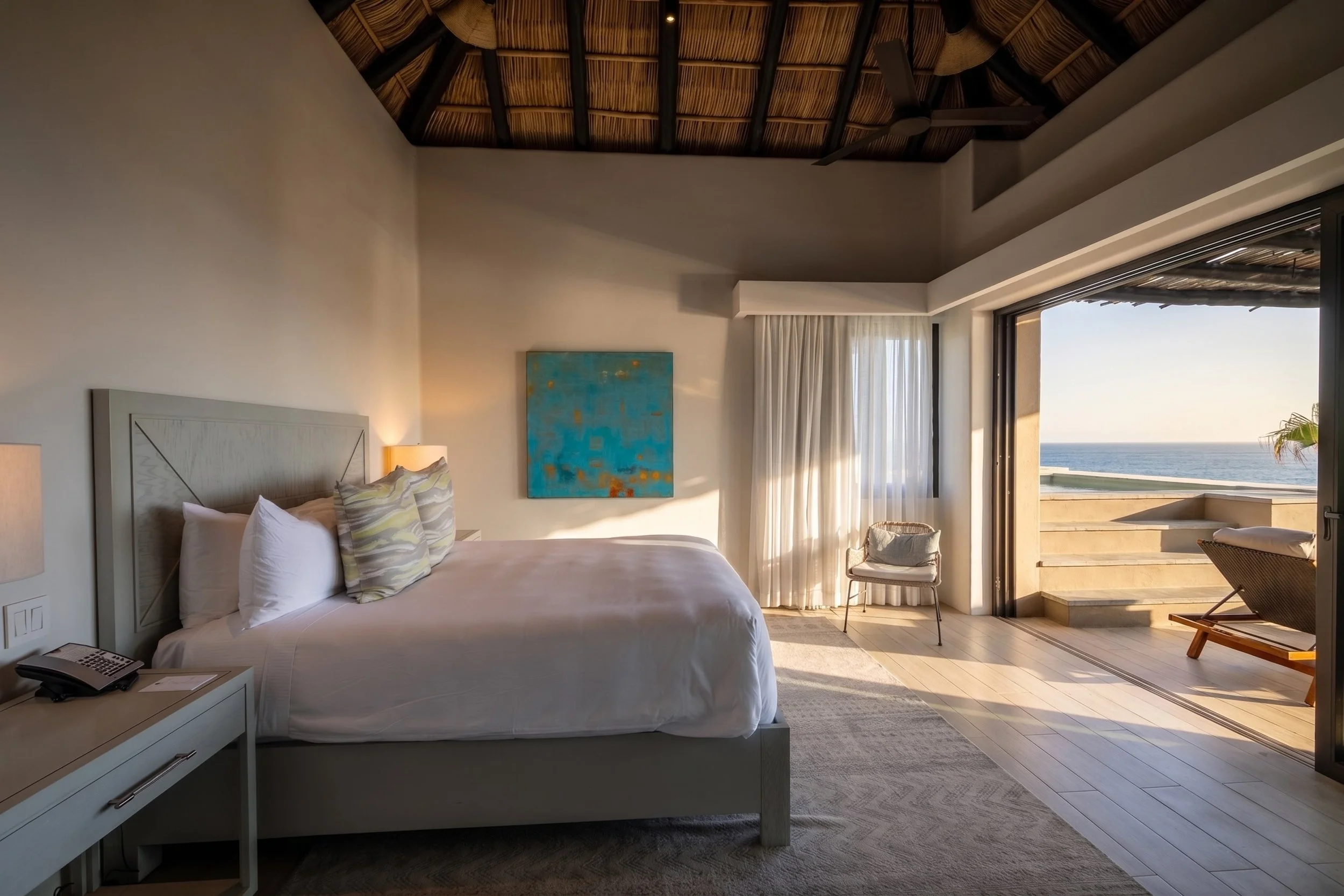 A bedroom with a large bed, artwork on the wall, a nightstand with a telephone, and a view of the ocean through an open sliding door leading to a balcony with stairs.