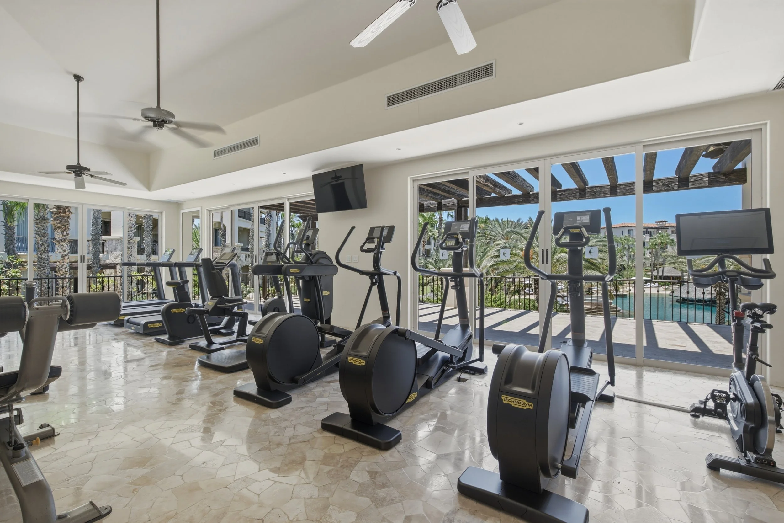 Indoor gym at Auburge Resort with exercise equipment such as ellipticals, stationary bikes, and treadmills, large windows showing outdoor patio and trees, ceiling fans, and a TV mounted on the wall.