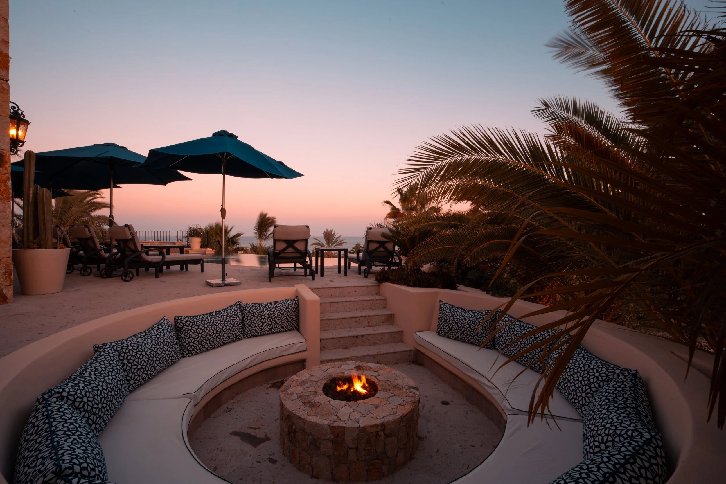 Outdoor seating area with a curved white couch with black patterned pillows, in front of a stone fire pit, overlooking a sunset view with palm trees, lounge chairs, and large umbrellas.