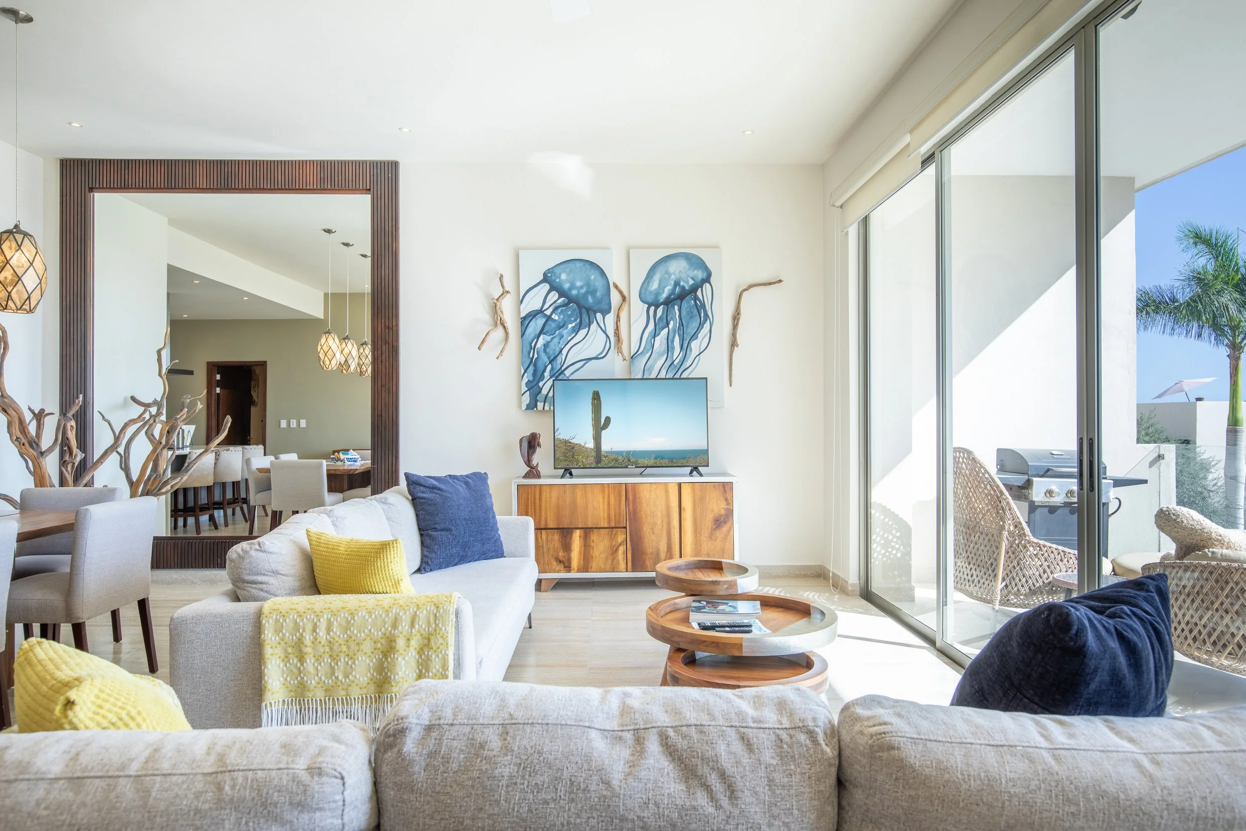 Inside a modern living room with white walls, a large window with sliding doors showing outdoor palm trees, a beige sofa with navy and yellow throw pillows, a wooden TV stand with a flat-screen TV, the wall decorated with jellyfish paintings, and a d