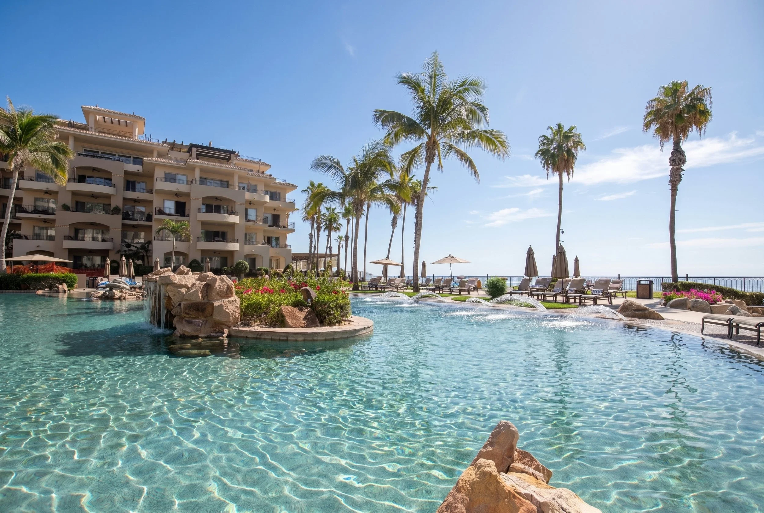 A resort with a swimming pool, palm trees, lounge chairs, and a multi-story building under a clear blue sky at Villa La Estancia