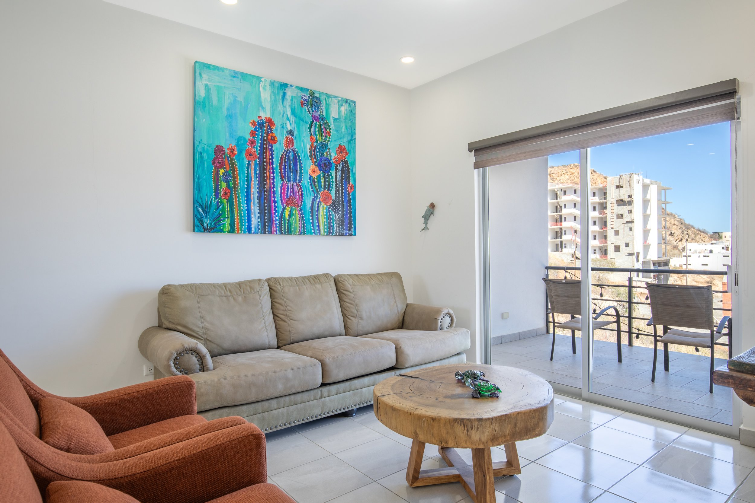 Living room with a beige sofa, a rust-colored armchair, a large wooden coffee table, and artwork of colorful cacti on the wall. There is a sliding glass door leading to a balcony with outdoor chairs, overlooking buildings and a hill outside.