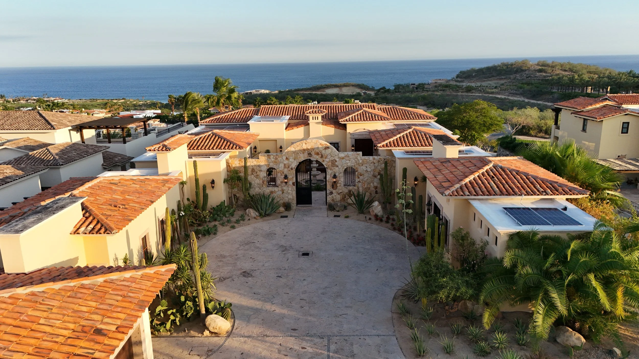 Luxury house with red tile roof, stone facade, desert landscaping including cacti, in front of a coastal view with blue ocean and hills in the background.