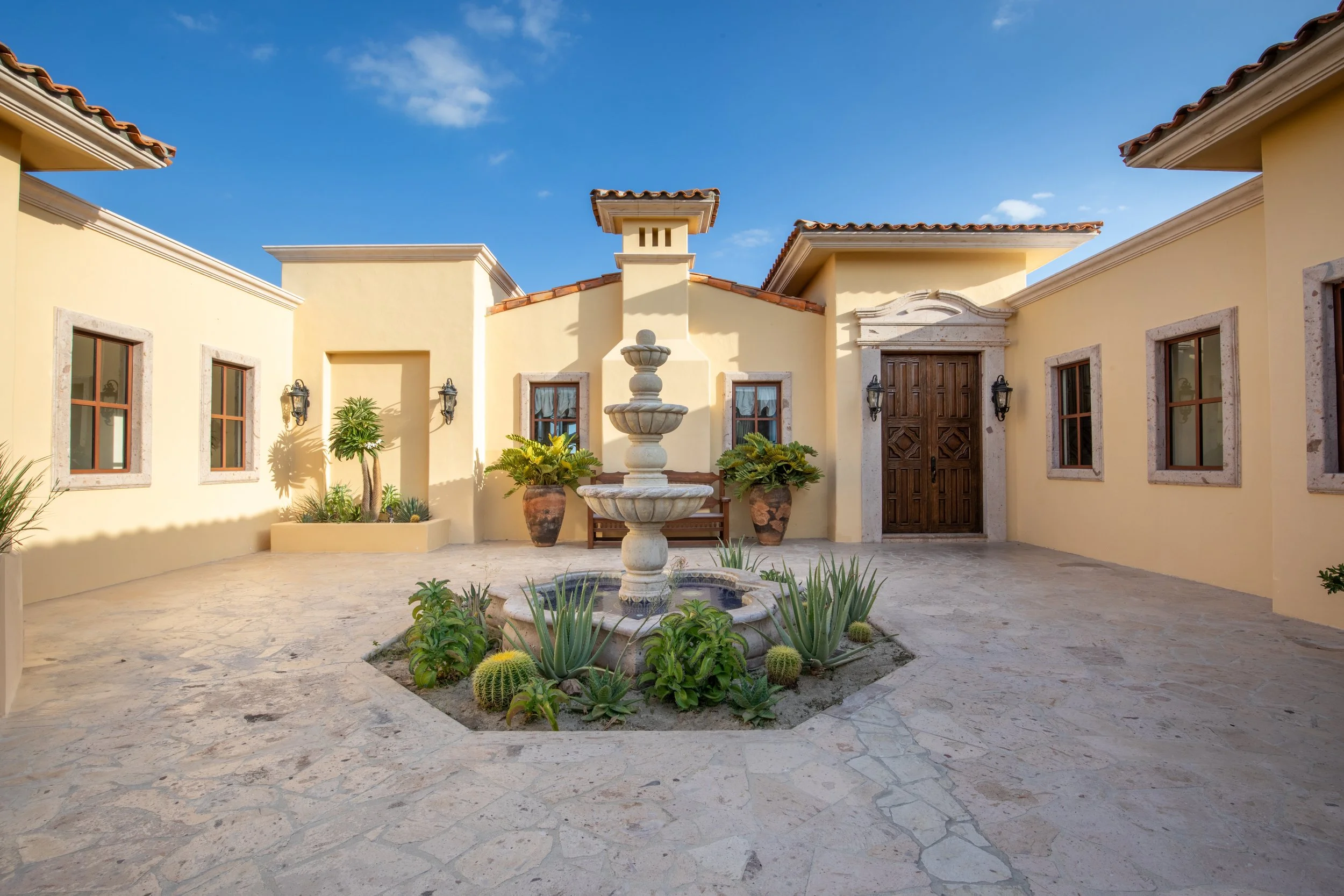 A Mediterranean-style courtyard with a water fountain in the center, surrounded by desert plants, potted greenery, and a yellow stucco building with wooden doors and windows.