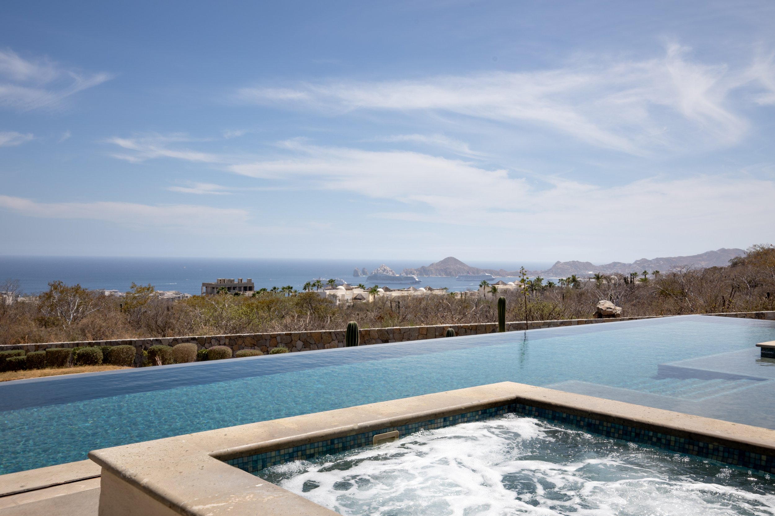 A swimming pool with a view of the ocean, mountains, and islands in the distance under a blue sky with some clouds. In Los Cabos Solaria residences.
