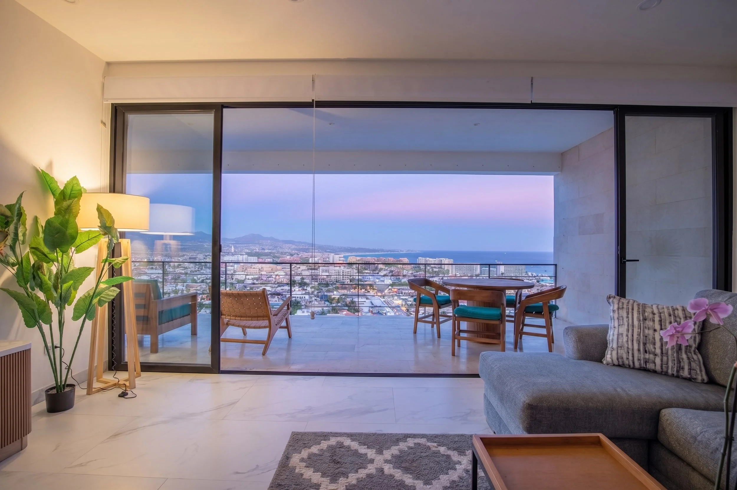 Living room in Montemar with a large glass sliding door opening to a balcony with a view of a city and mountains. The room has a grey sofa with a striped pillow, a coffee table, a potted plant, a standing lamp, and chairs around a small dining table.