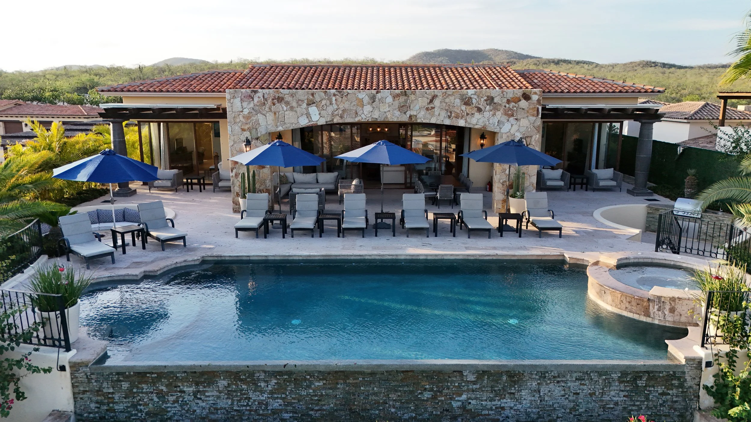 Luxury house with a swimming pool, sun loungers, and blue umbrellas in a backyard, with hills in the background.