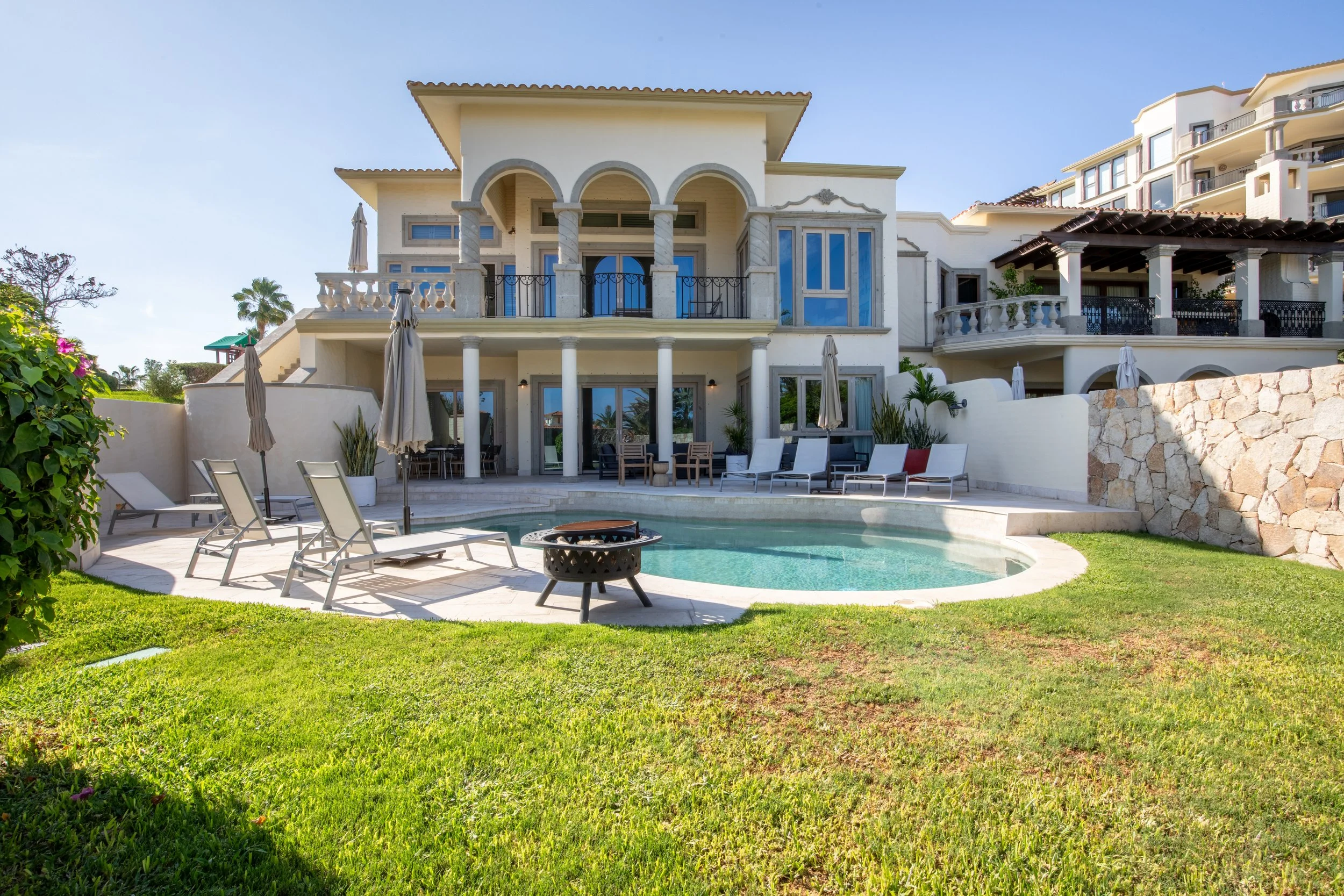 A luxurious house with a swimming pool in the front yard, surrounded by green grass and lounge chairs, with multiple balconies and large windows, under a clear blue sky.