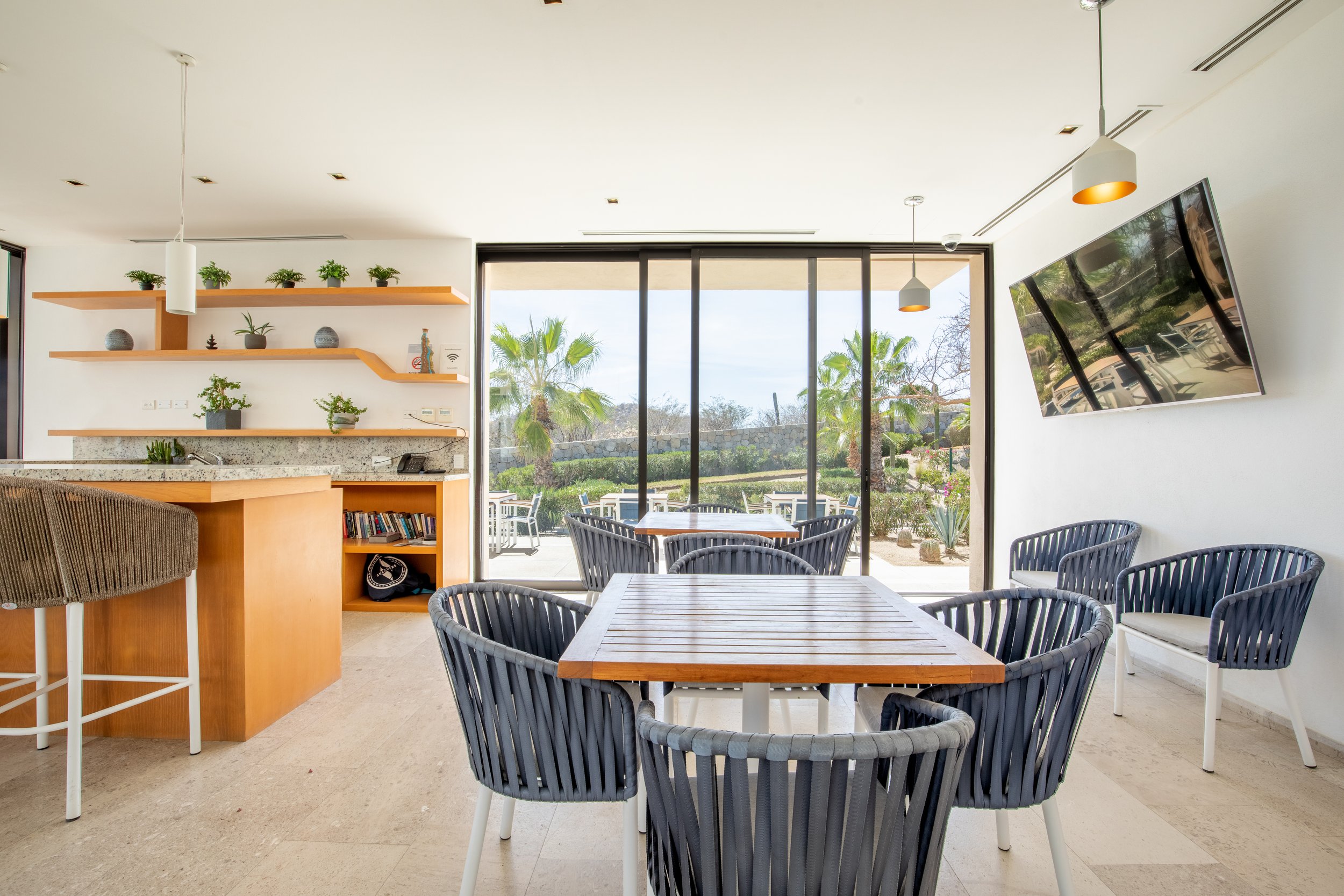 Modern indoor dining area with wooden tables, gray woven chairs, and large glass sliding doors opening to an outdoor patio with palm trees and greenery.