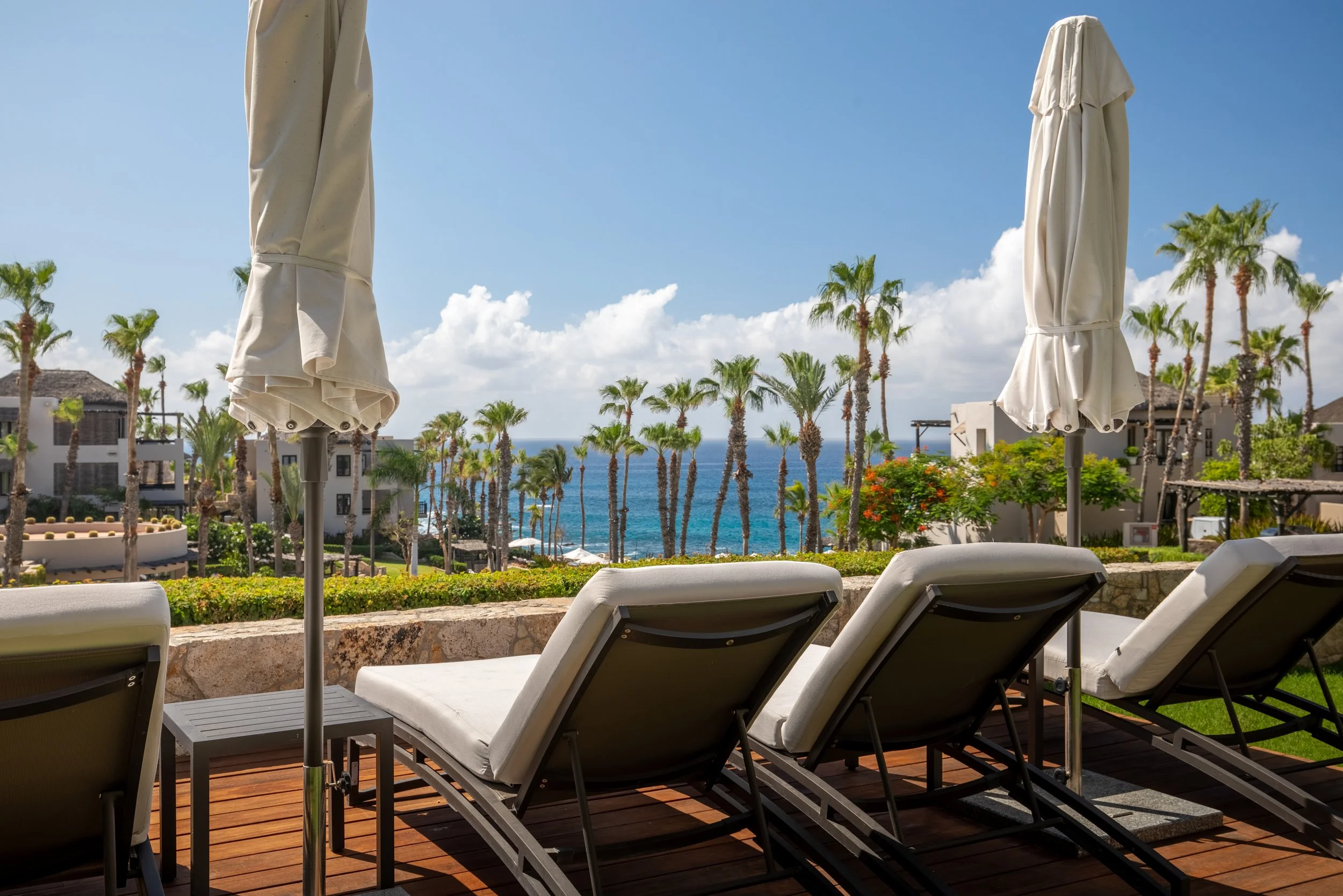Poolside with lounge chairs and umbrellas overlooking a tropical landscape with palm trees and buildings, ocean view in the background on a sunny day.