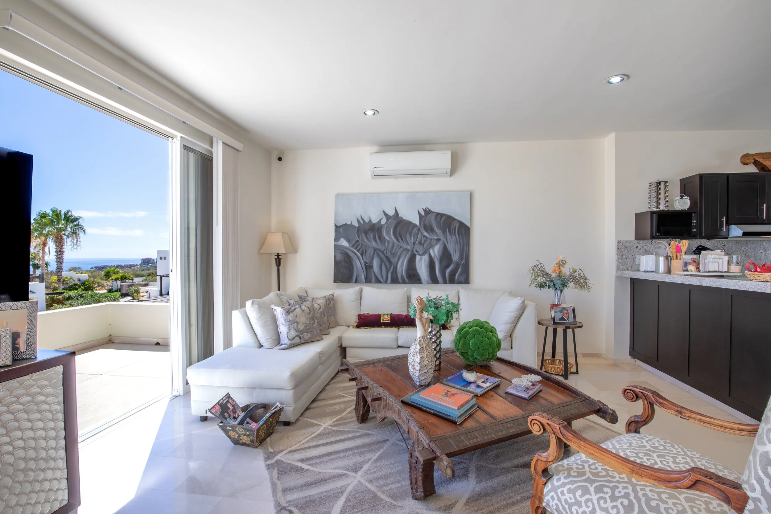 A bright living room with white sofa, dark wooden coffee table, and artwork of black horses on the wall. Large sliding glass door opens to an outdoor patio with palm trees and ocean view.