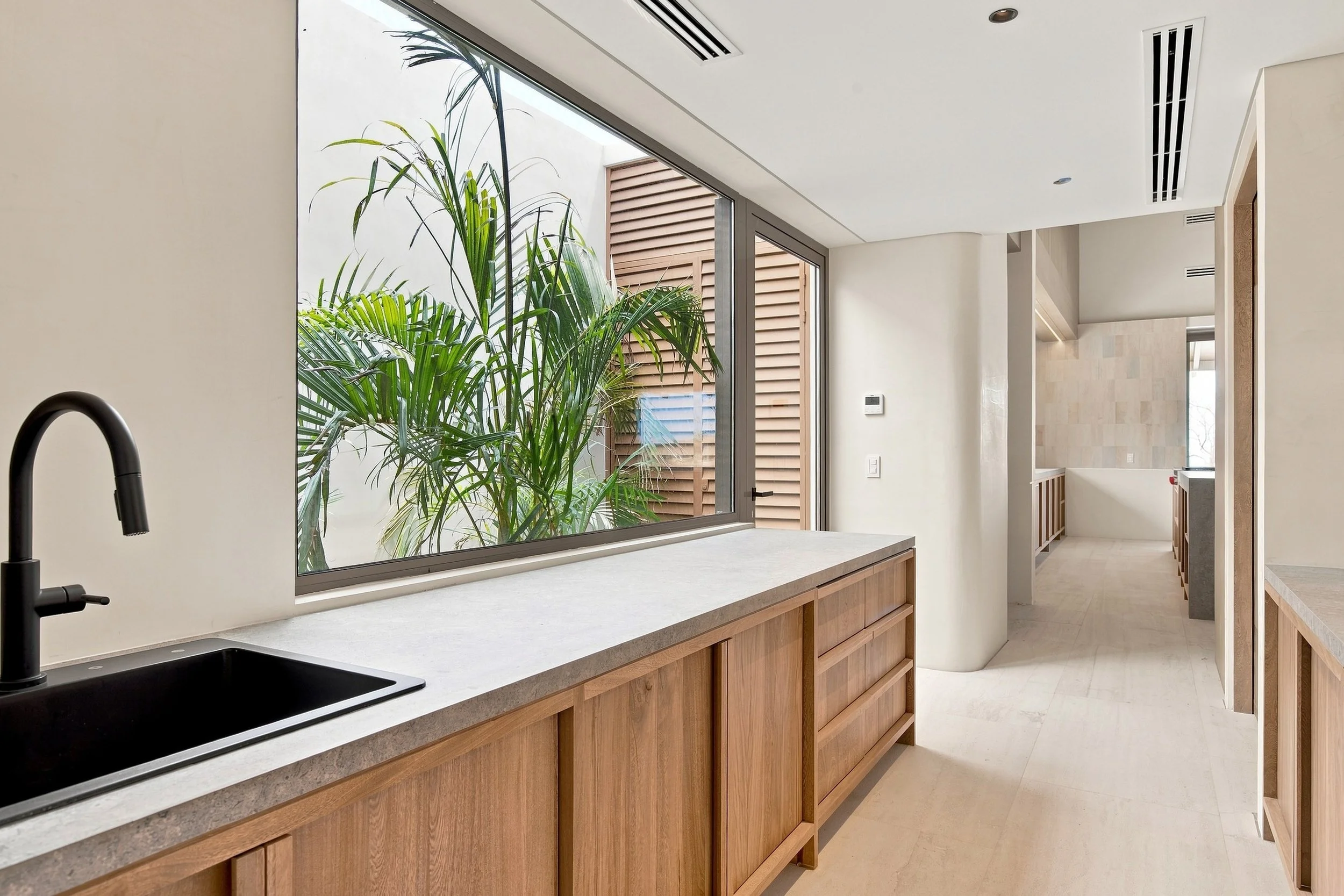 Modern kitchen with large window, black sink faucet, white countertops, wooden cabinets, and a leafy plant outside the window.