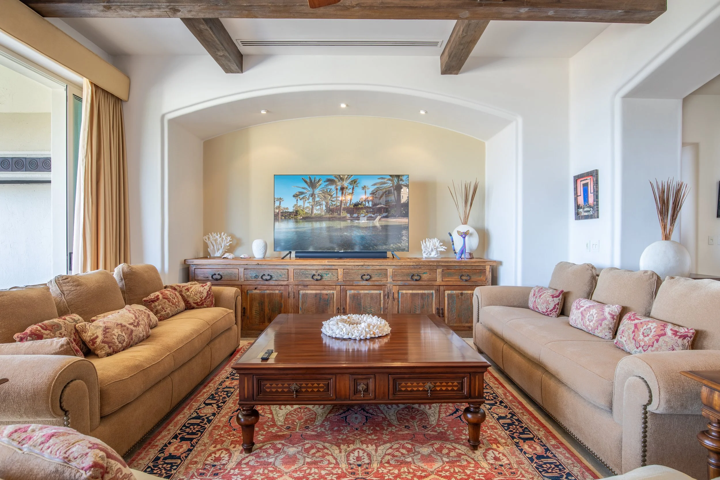 Living room with two beige sofas, a wooden coffee table with ceramic sea shells decor, a large flat-screen TV on a wooden console, art and decorative vases with dried plants, adjacent to a sliding glass door with beige curtains, and a patterned area 