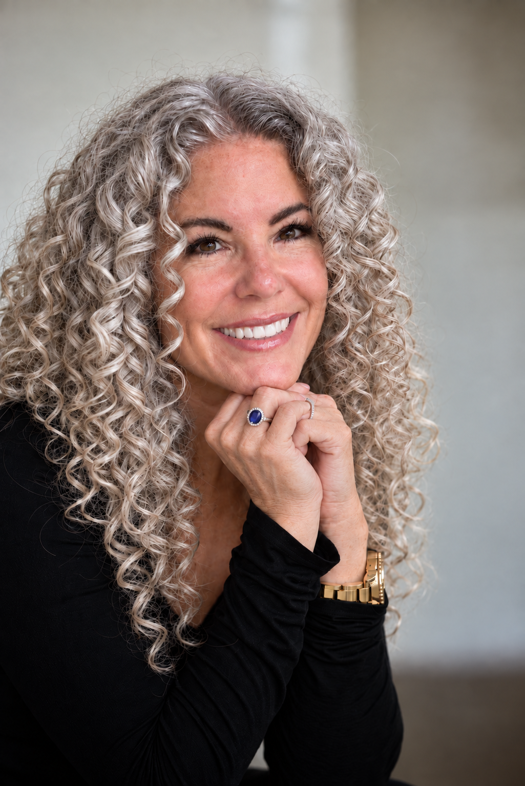 A woman with long, curly gray hair smiling at the camera, wearing a black top, a blue ring, a gold watch, and a delicate ring on her finger, resting her chin on her hands.