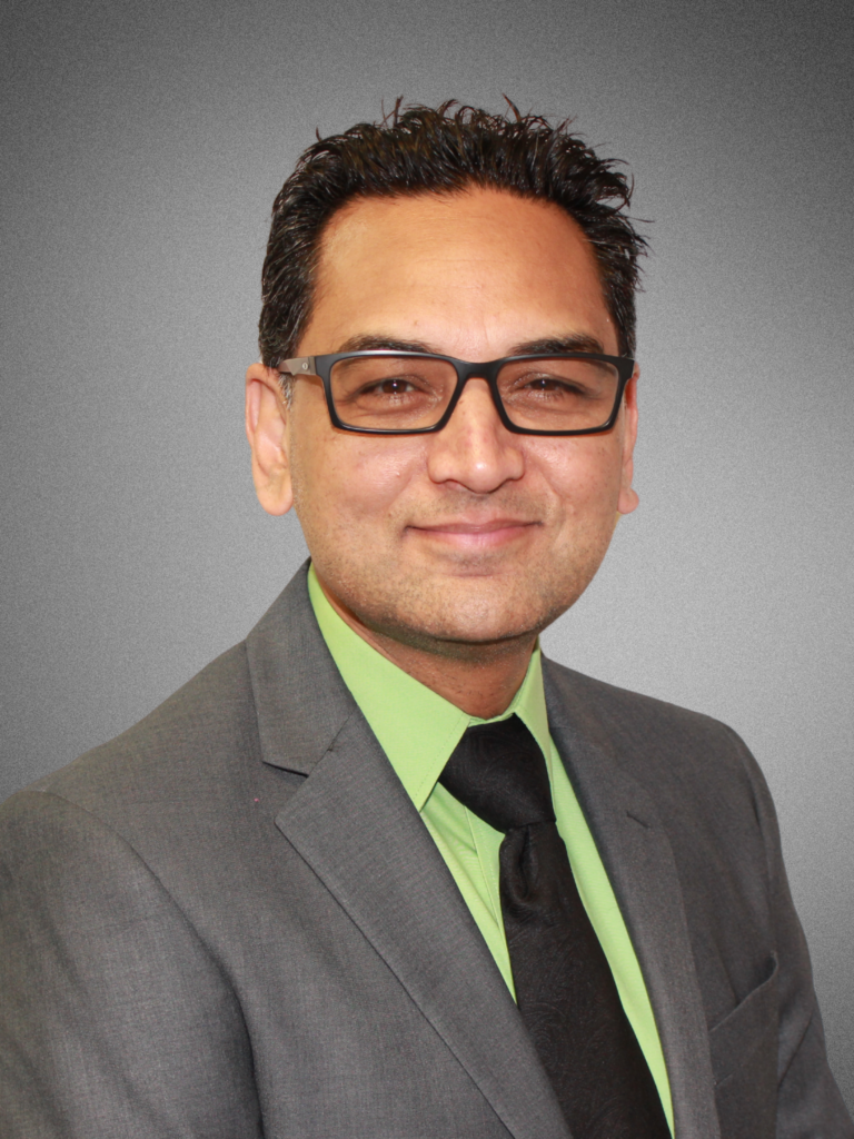 Portrait of a man wearing glasses, a grey suit, a lime green shirt, and a black tie, smiling slightly against a plain grey background.