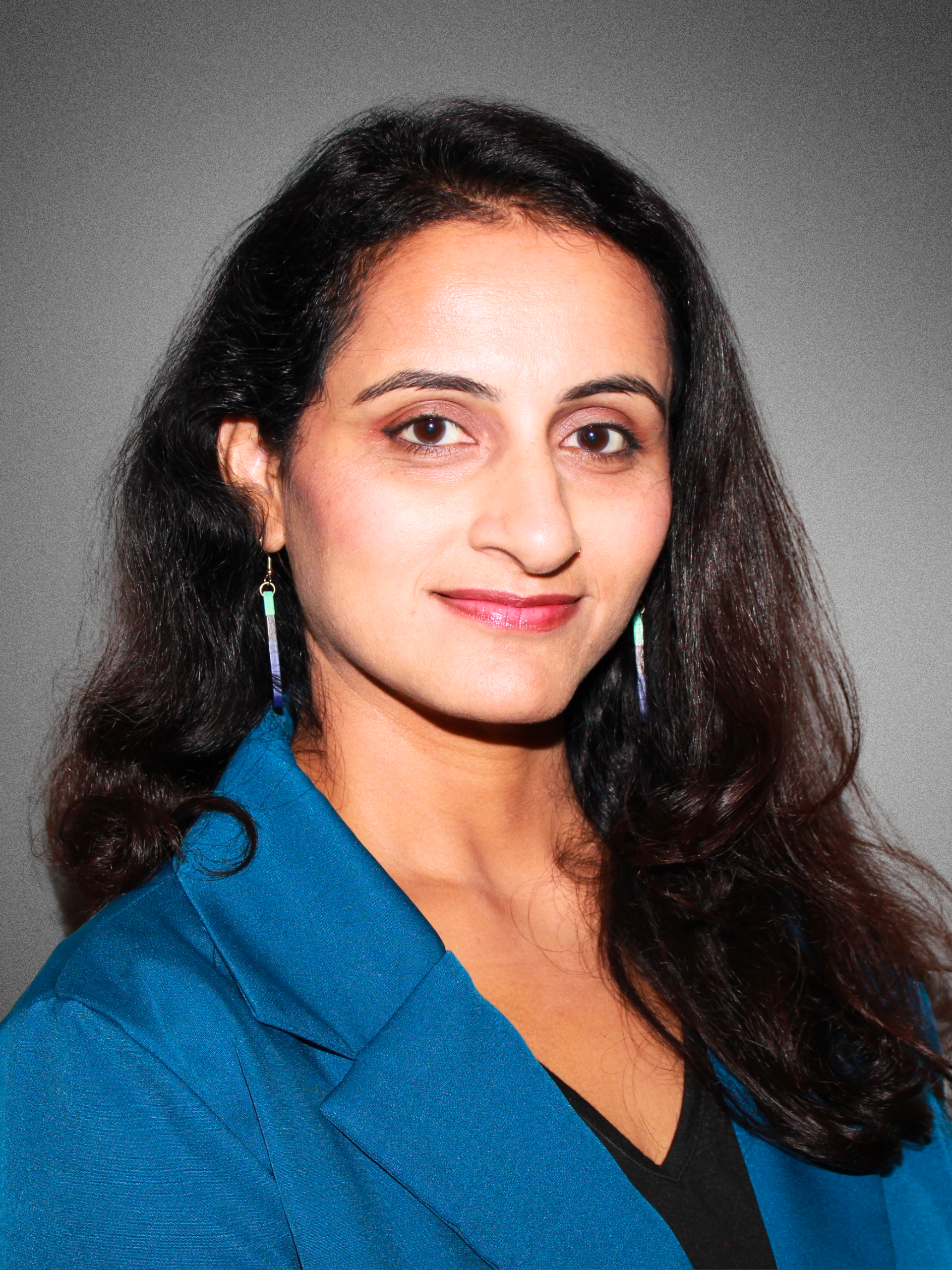 A woman with long dark wavy hair wearing a blue blazer, earrings, and a black top, smiling softly at the camera against a gray background.