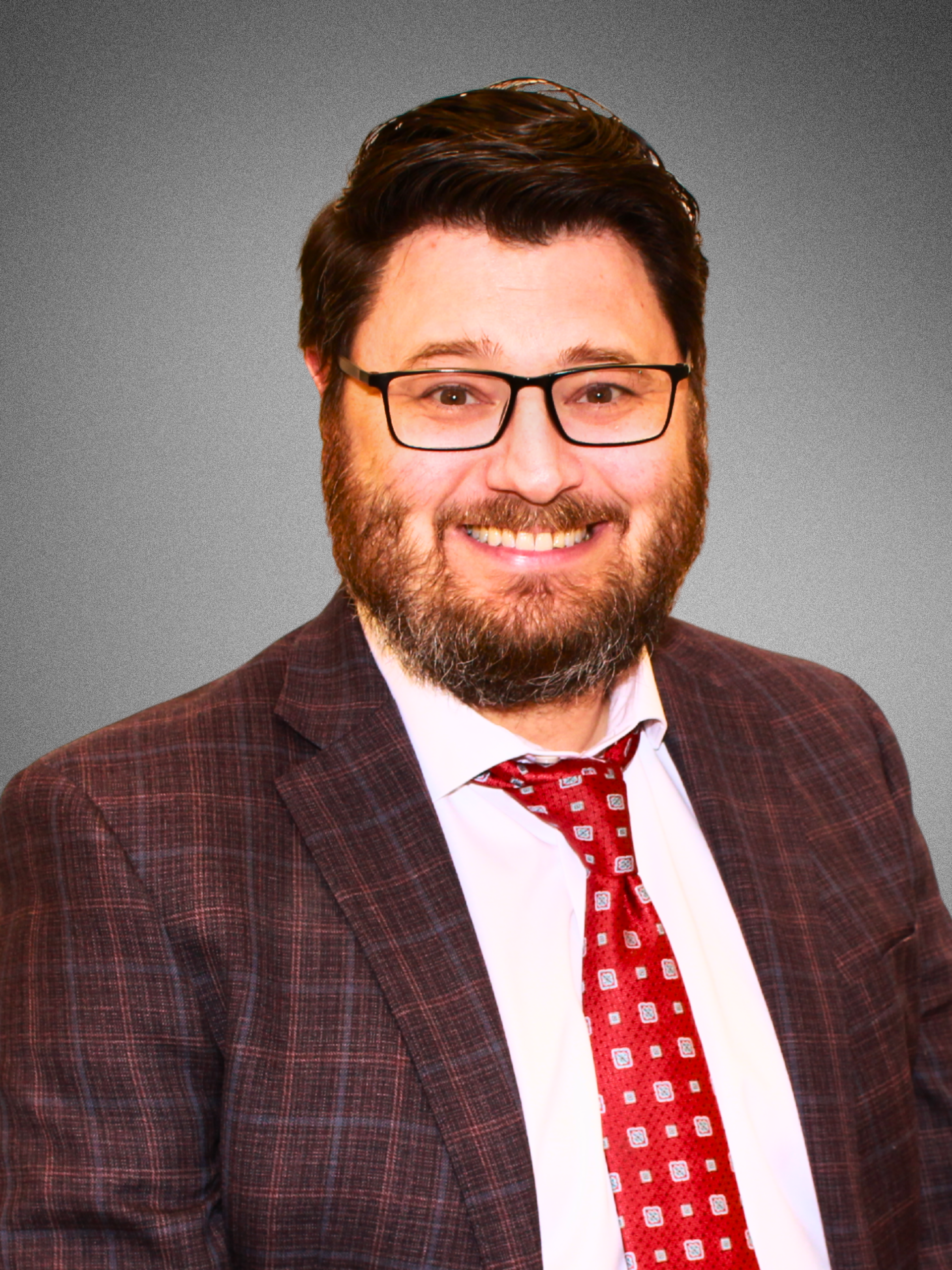 Headshot of a man with dark hair, wearing a checkered blue shirt, pink tie, and plaid suit jacket, smiling against a gray background.