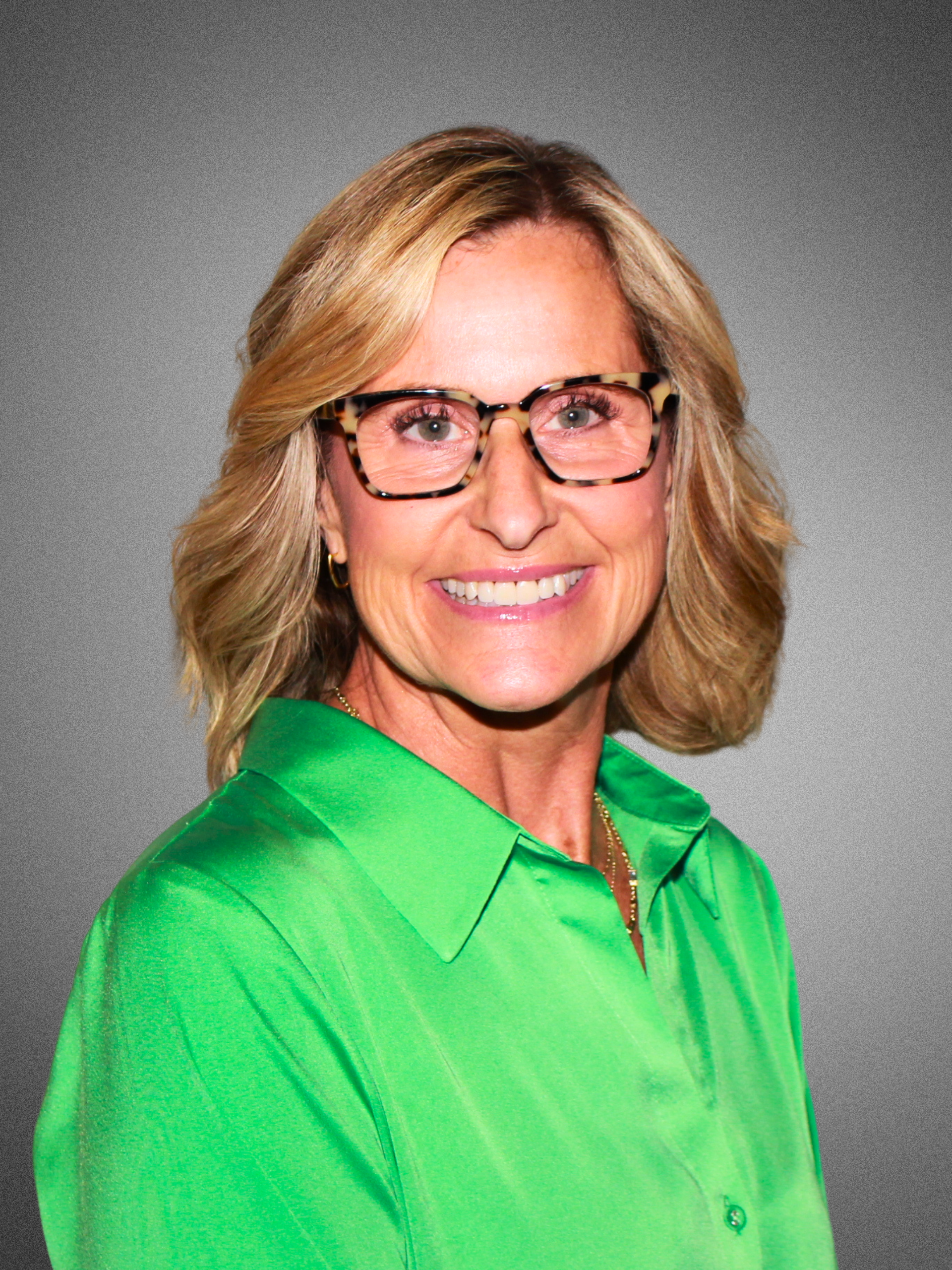 Portrait of a middle-aged woman with blonde hair, wearing glasses with leopard print frames, and a bright green blouse, smiling against a gray background.