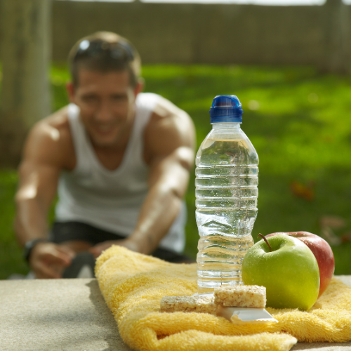 A young man smiling outdoors on a sunny day, with a water bottle, apples, a towel, and a snack in the foreground.