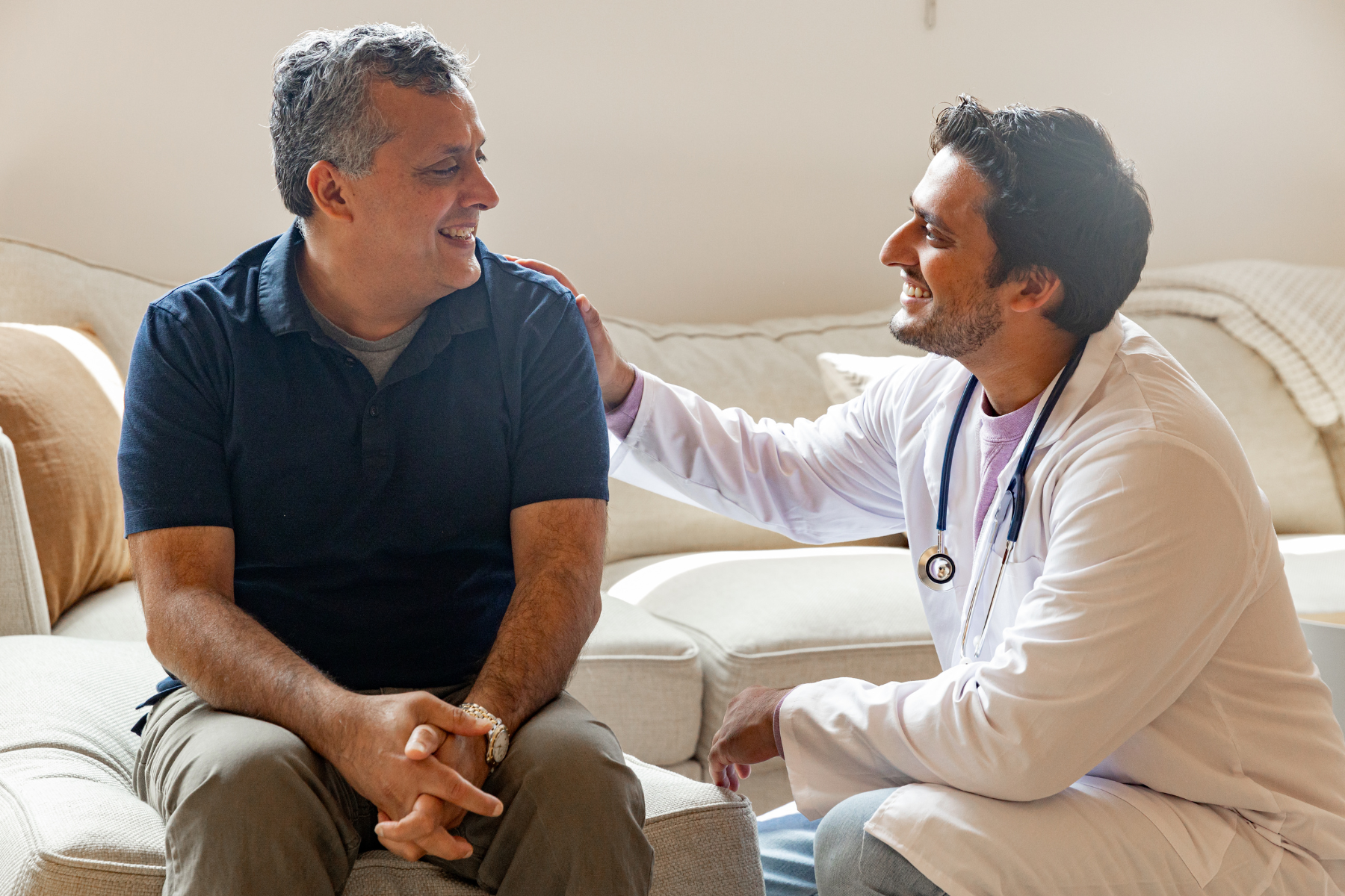 A healthcare professional, wearing a white coat and stethoscope, comforting a middle-aged man sitting on a beige couch in a well-lit room.