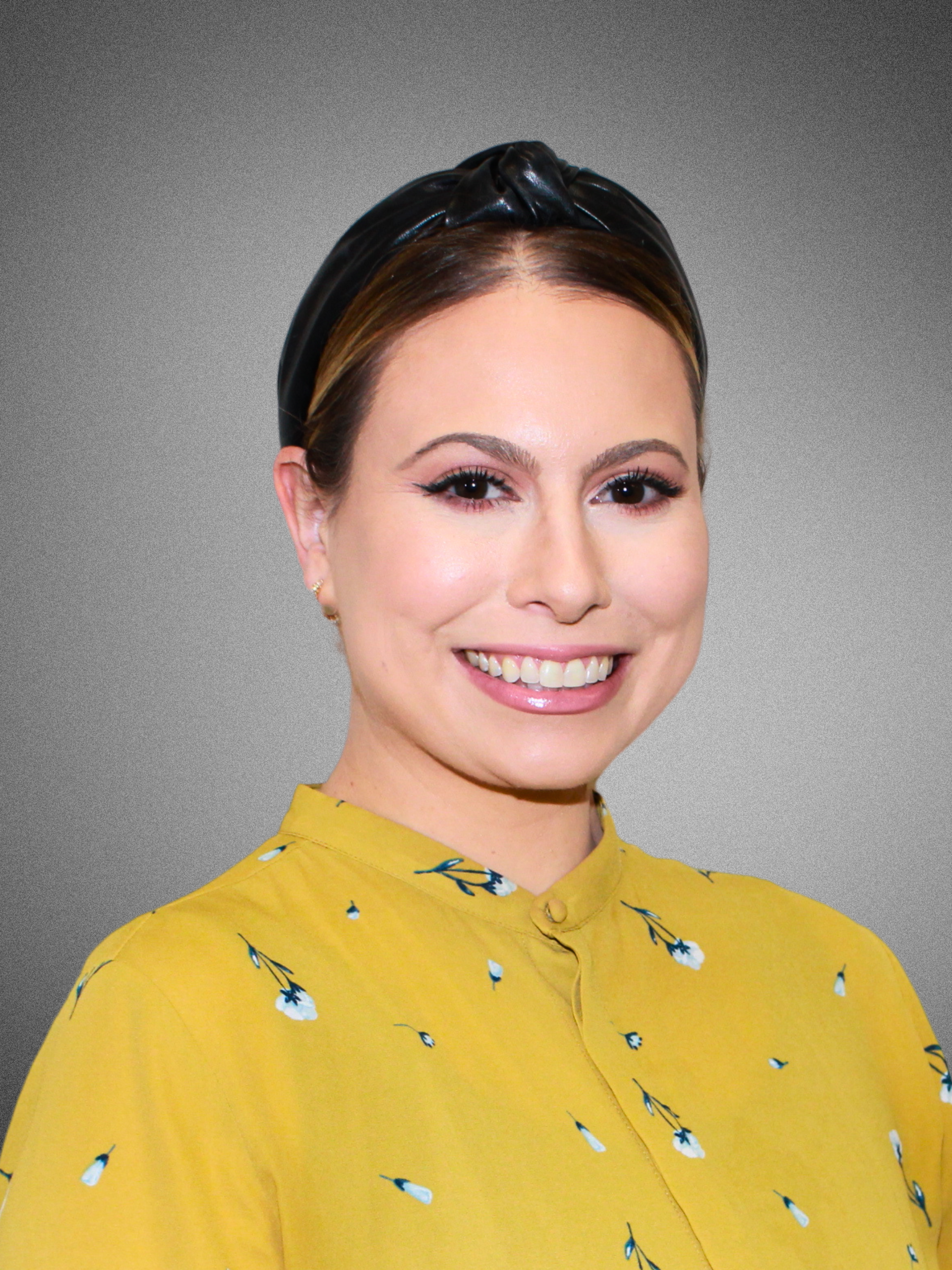 A smiling woman with fair skin, wearing a black headband and yellow blouse with a floral pattern, against a gray background.