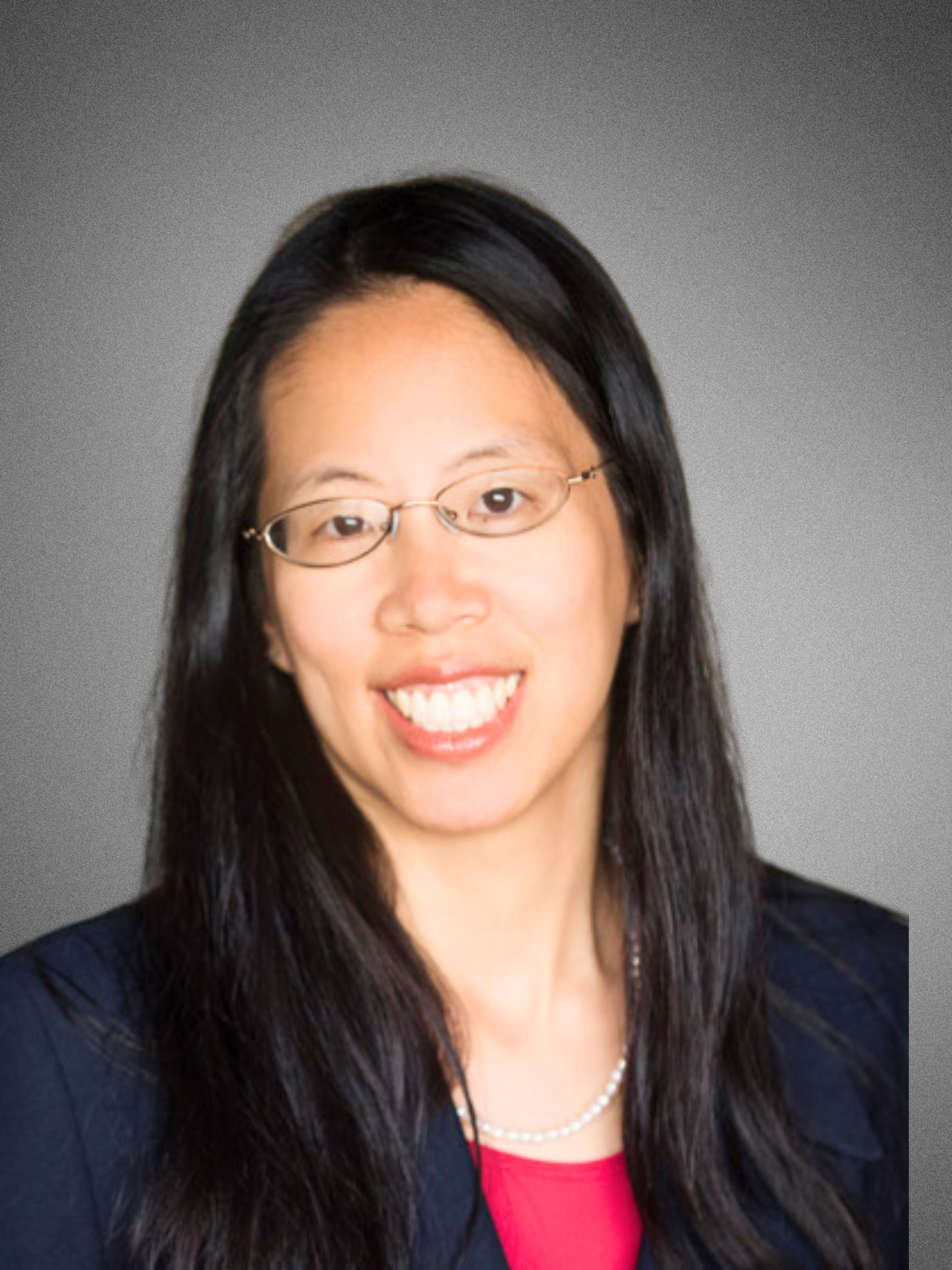 Professional headshot of a woman with long black hair, glasses, wearing a dark blazer, pink top, and pearl necklace, smiling against a gradient gray background.