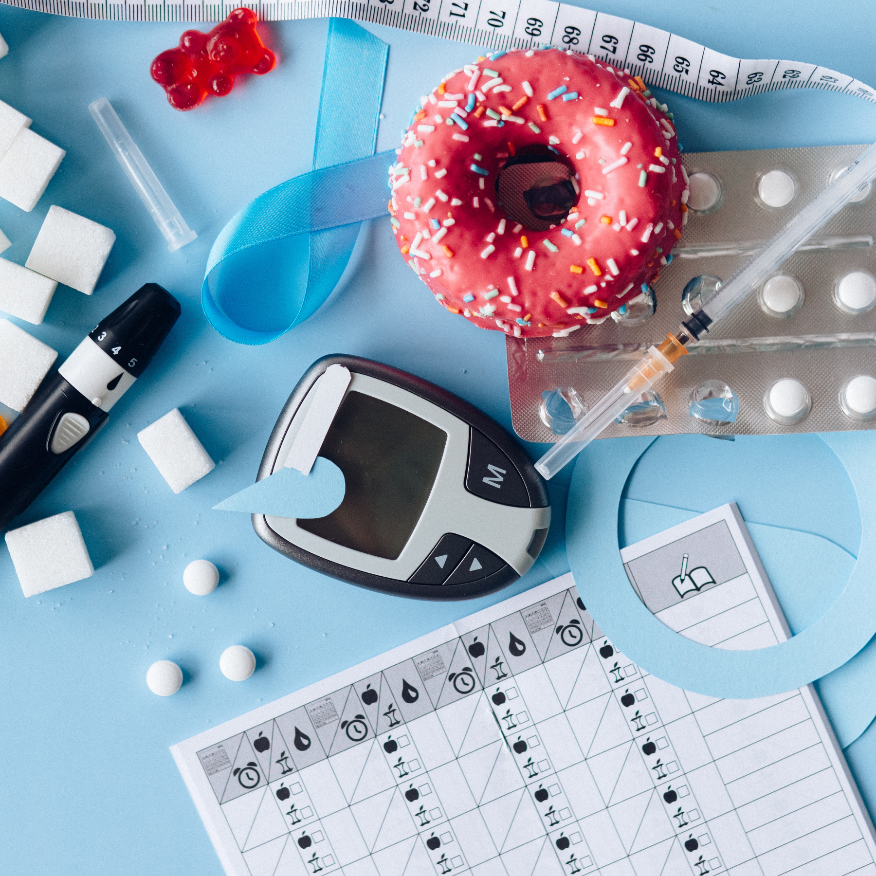 A diabetic test kit includes a blood glucose monitor, test strips, and lancets, surrounded by sugar cubes, a pink sprinkled donut, a measuring tape, insulin syringe, and diabetes management charts, all on a blue background.