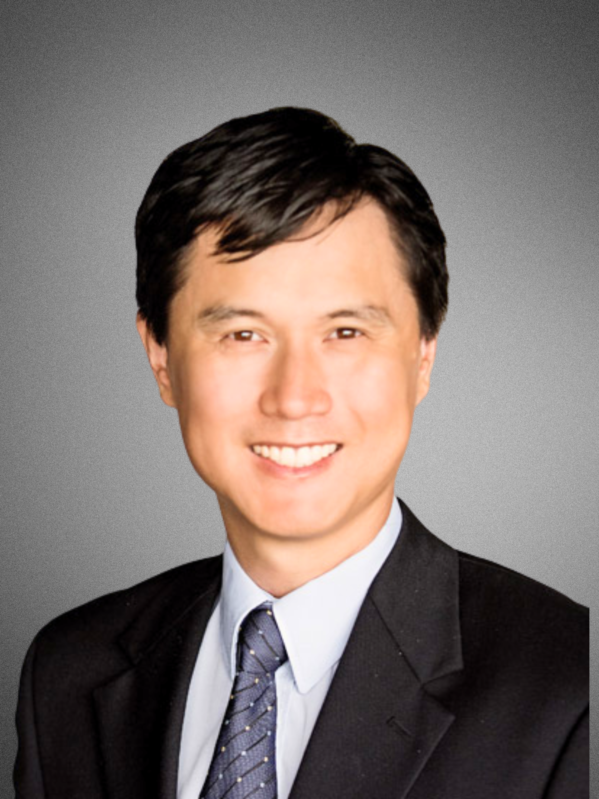 Professional headshot of a smiling man with dark hair in a suit and tie against a grey background.