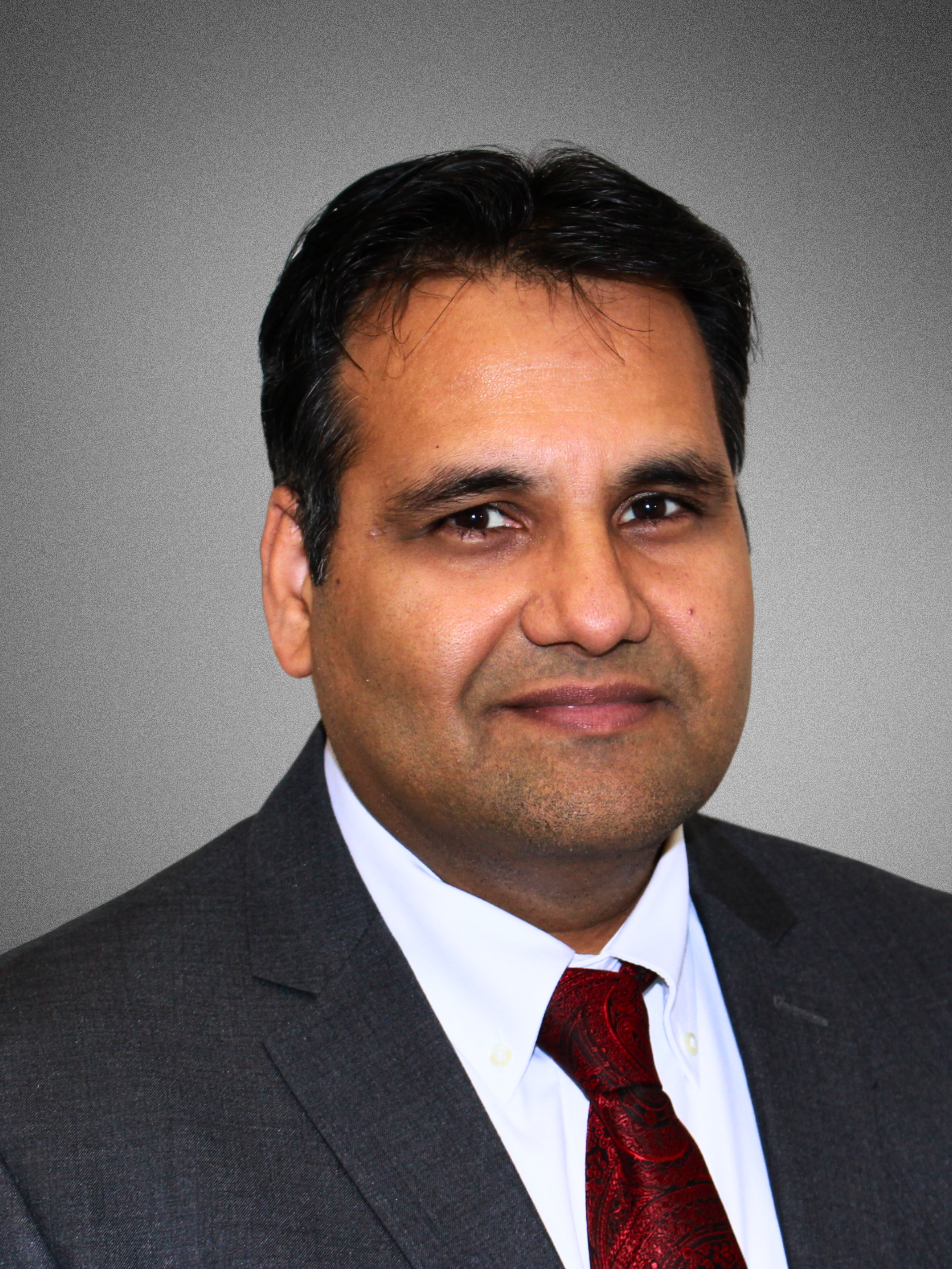 Professional headshot of a man in a dark suit, white shirt, and red tie, with a neutral gray background.