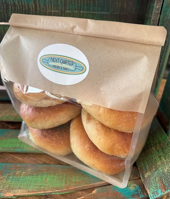 Bag of bread rolls with a sticker labeled "Next Chapter Breads & Bakes" on a wooden surface.