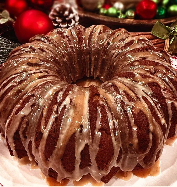 A bundt cake topped with glaze snowing over it, with Christmas decorations like a red ornament, pine cone, and green and red baubles in the background.