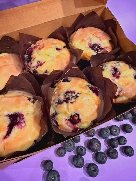 Six blueberry muffins in a cardboard box with dark paper liners, surrounded by scattered blueberries on a purple surface.