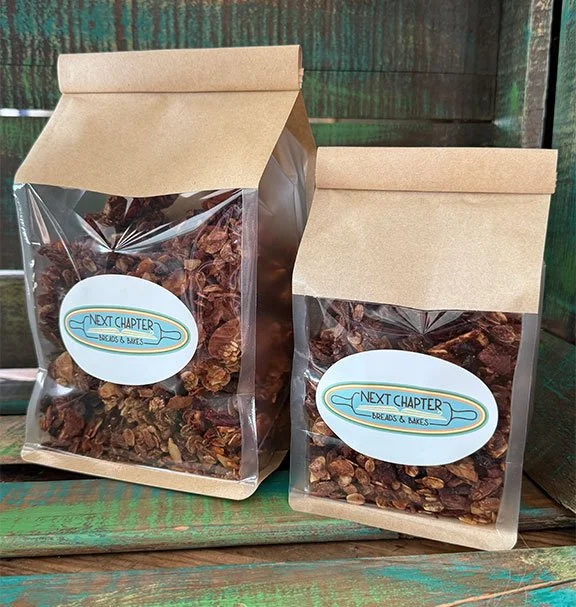Two resealable brown paper bags filled with granola, sitting on a weathered green wooden surface, with a logo sticker reading "Next Chapter Breaks & Bakes."
