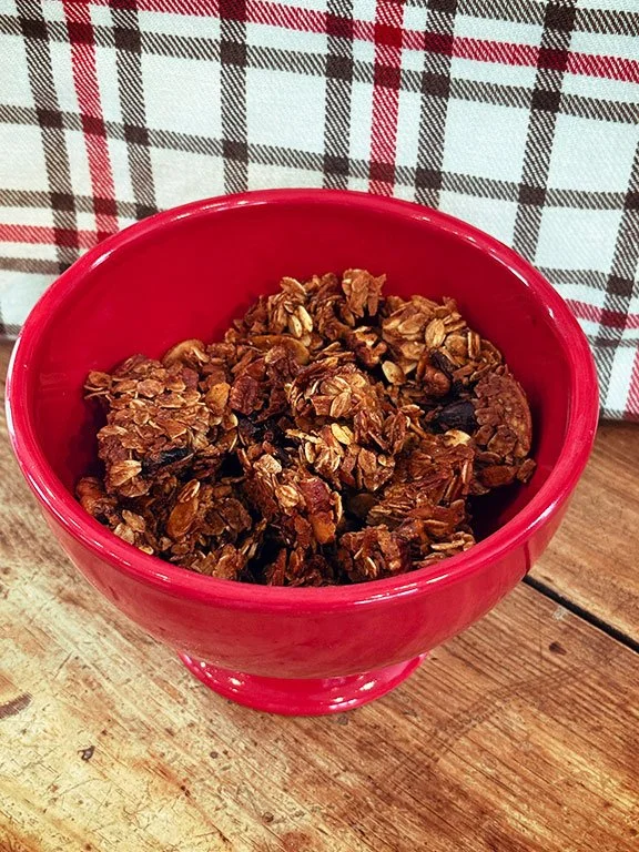 Granola in a red bowl on a wooden table with a checked cloth in the background.