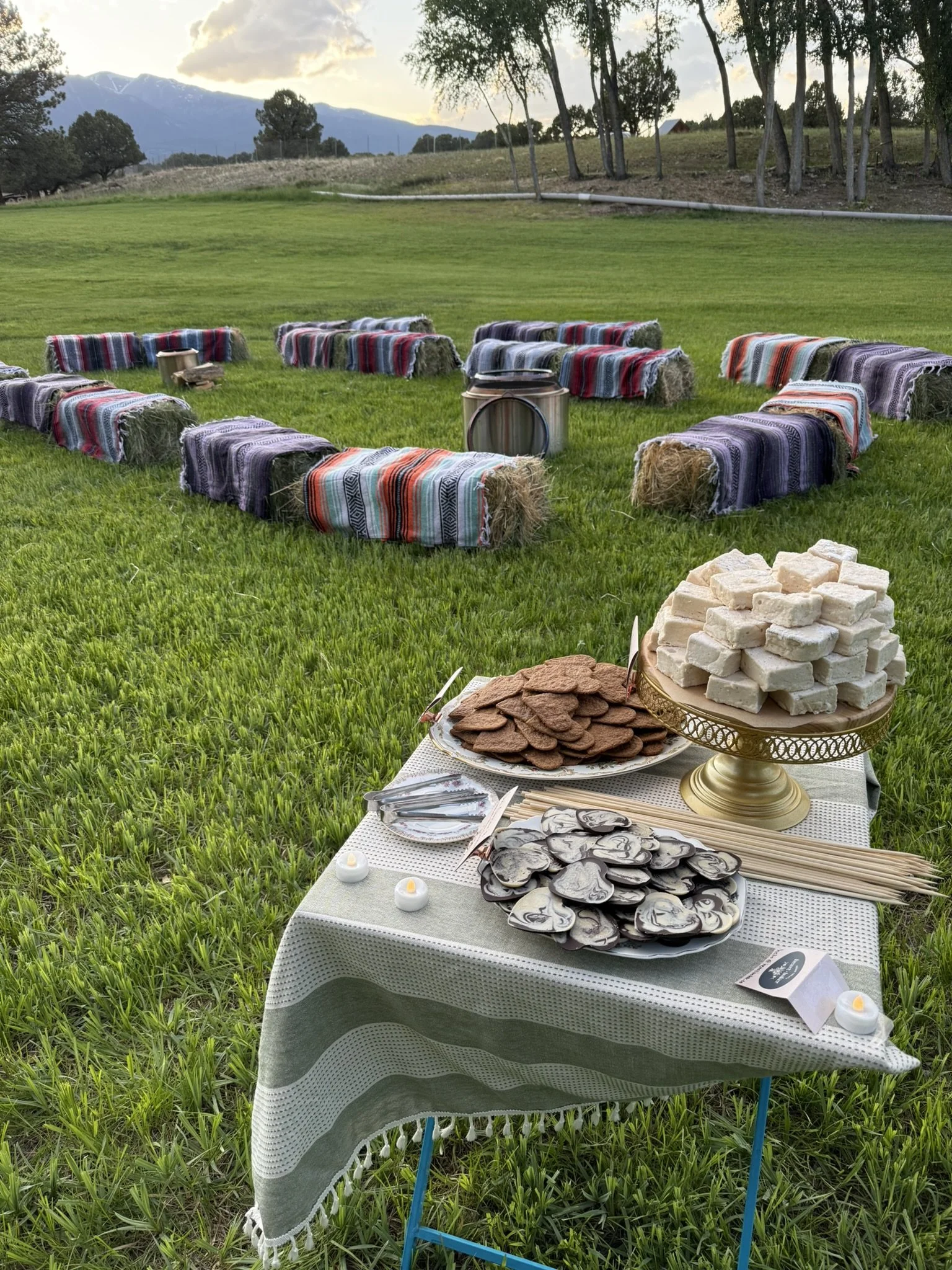 A rustic outdoor gathering setup with hay bale seating covered in colorful blankets, arranged in a circle on a grassy field. In the foreground, a table is decorated with a white tablecloth, candles, and plates of cookies, marshmallows, and chocolate 
