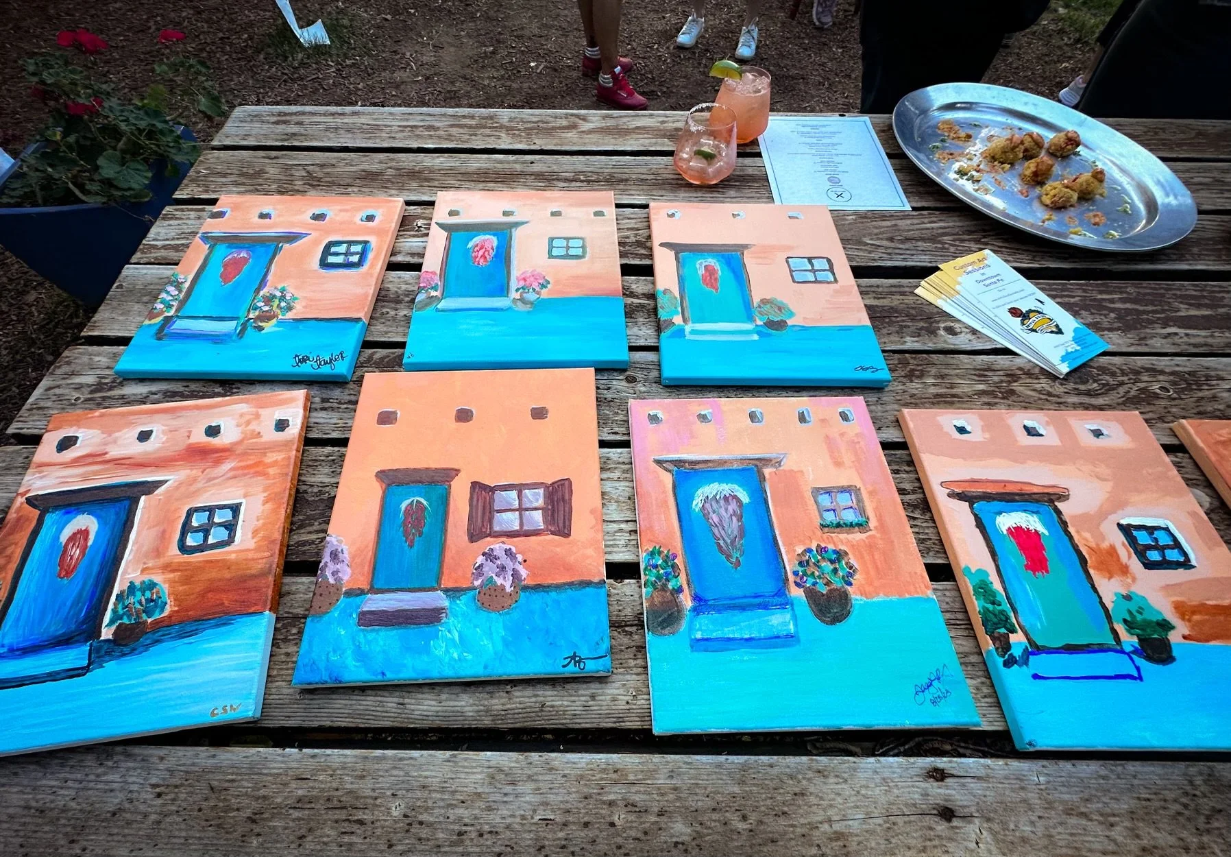 Paintings of doors and windows on small canvases arranged on a wooden table outdoors.