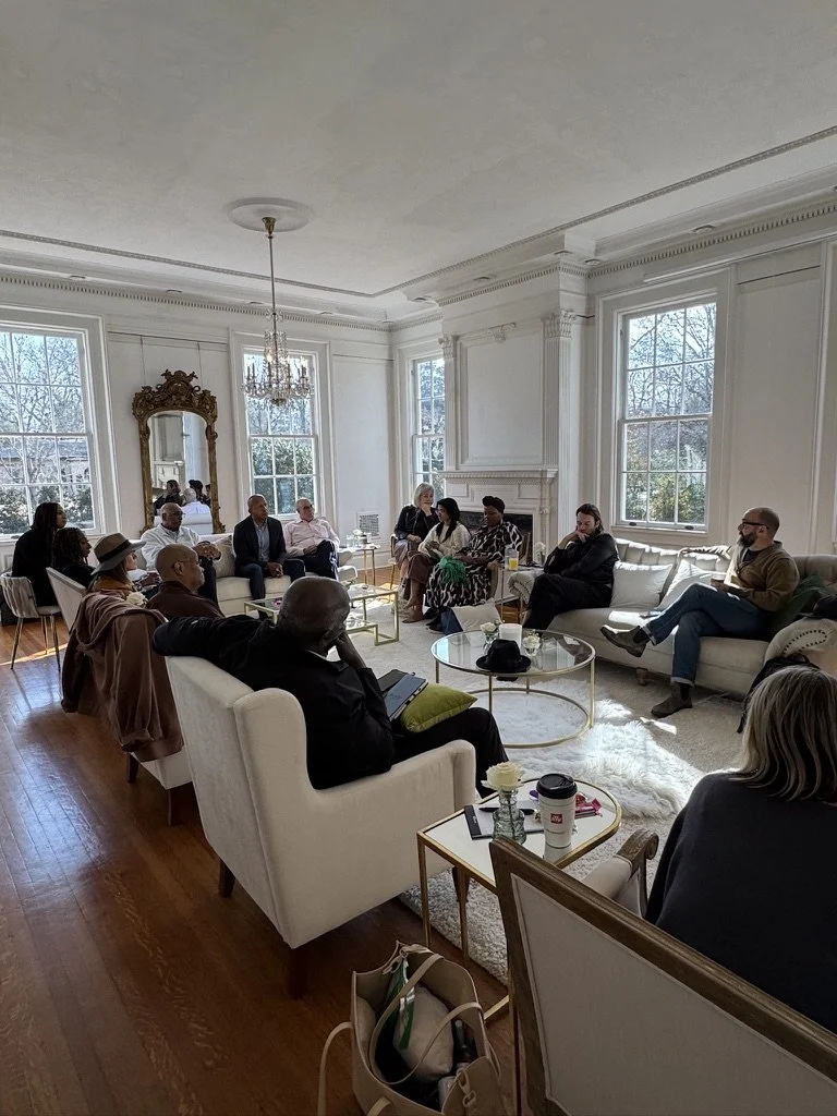 A group of people gathered in a bright, elegant living room with white walls, large windows, and a chandelier. They are seated on sofas and chairs, engaged in a discussion.