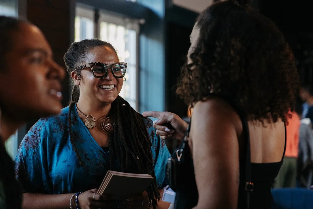 Three women engaged in conversation at an indoor event, smiling and gesturing, with the woman in the center holding a notebook.