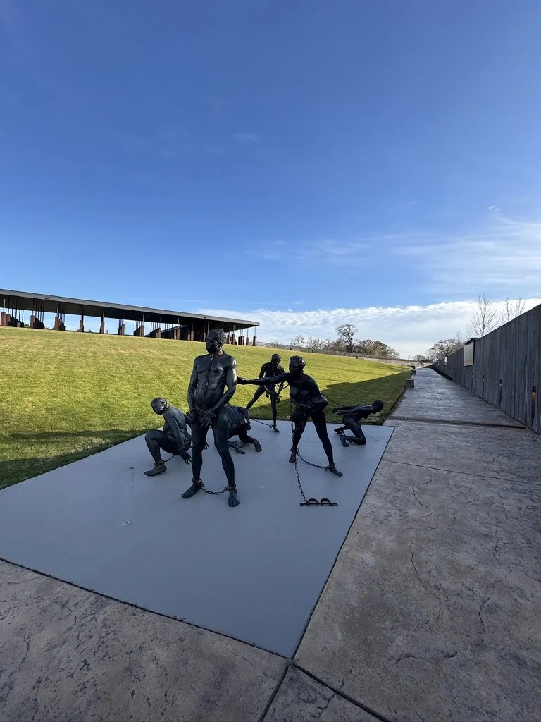 Statue sculpture of men in chains, with some men in poses of struggle and others helping each other, on a concrete platform outdoors against a grassy hill, clear blue sky.