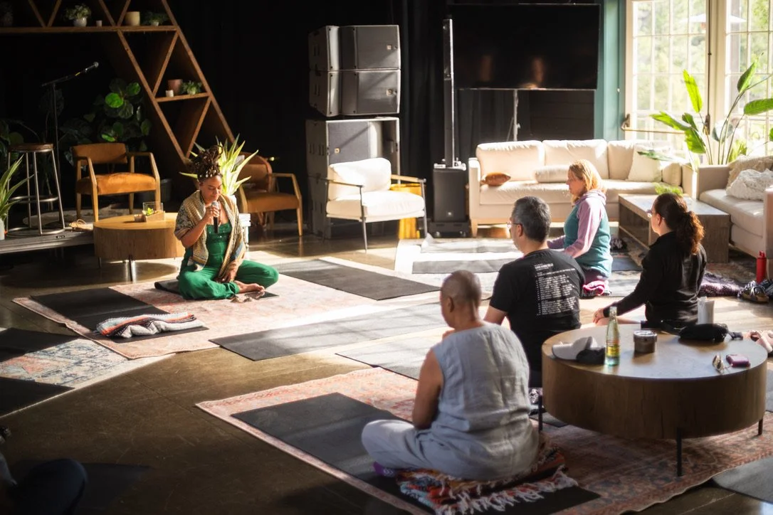 A group of five people participating in a meditation or wellness session in a cozy, well-lit room with plants, rugs, couches, and large windows. A woman in a colorful outfit is leading the session while the others sit cross-legged on mats.