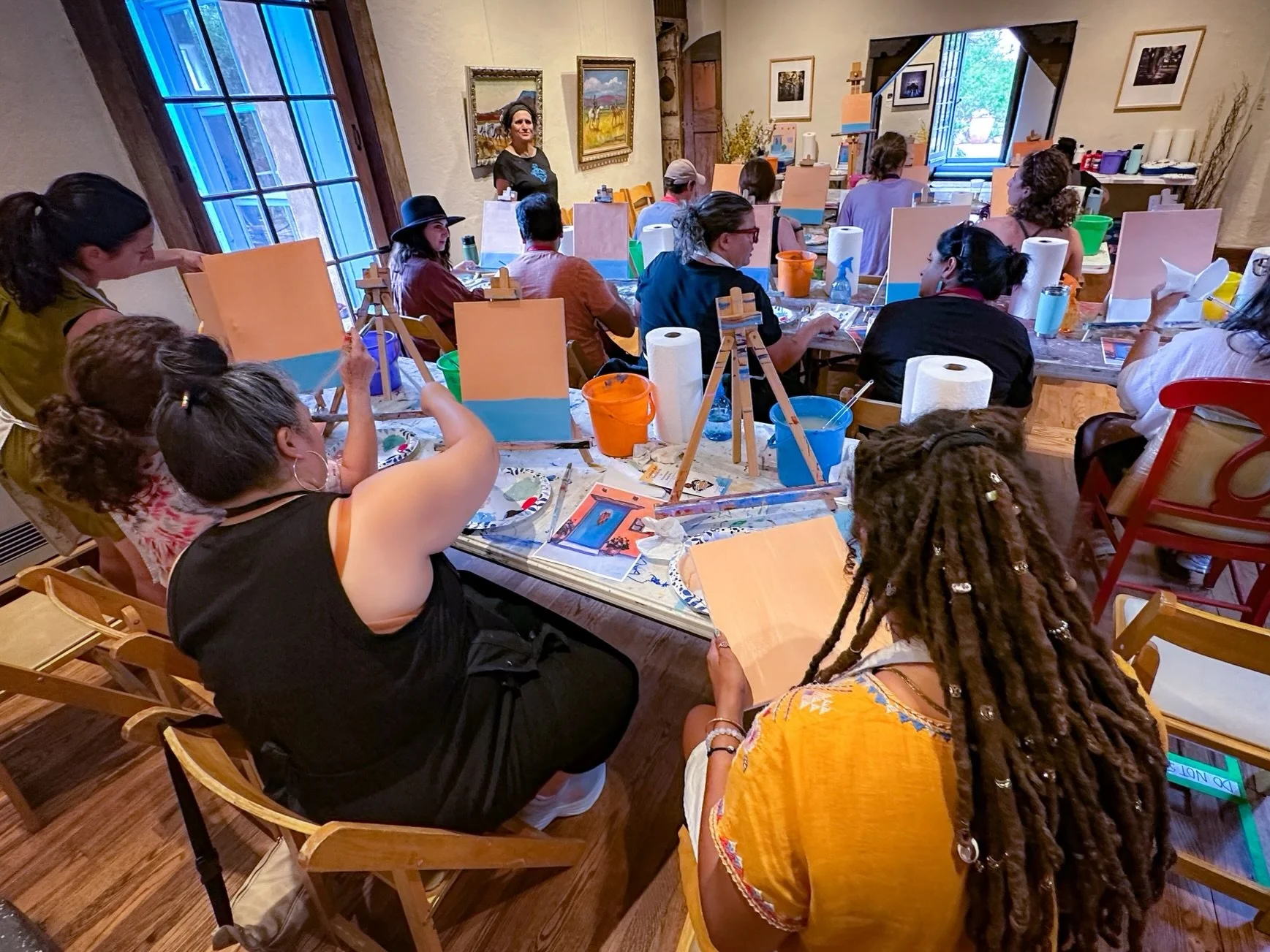Group of people participating in a painting class in a cozy indoor studio with paintings on the walls and large windows.