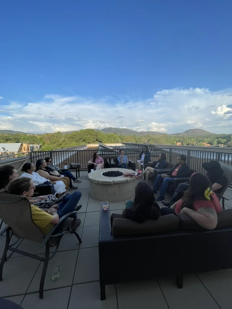 People sitting in a semi-circle on a rooftop terrace around a fire pit, with mountains and a blue sky in the background.