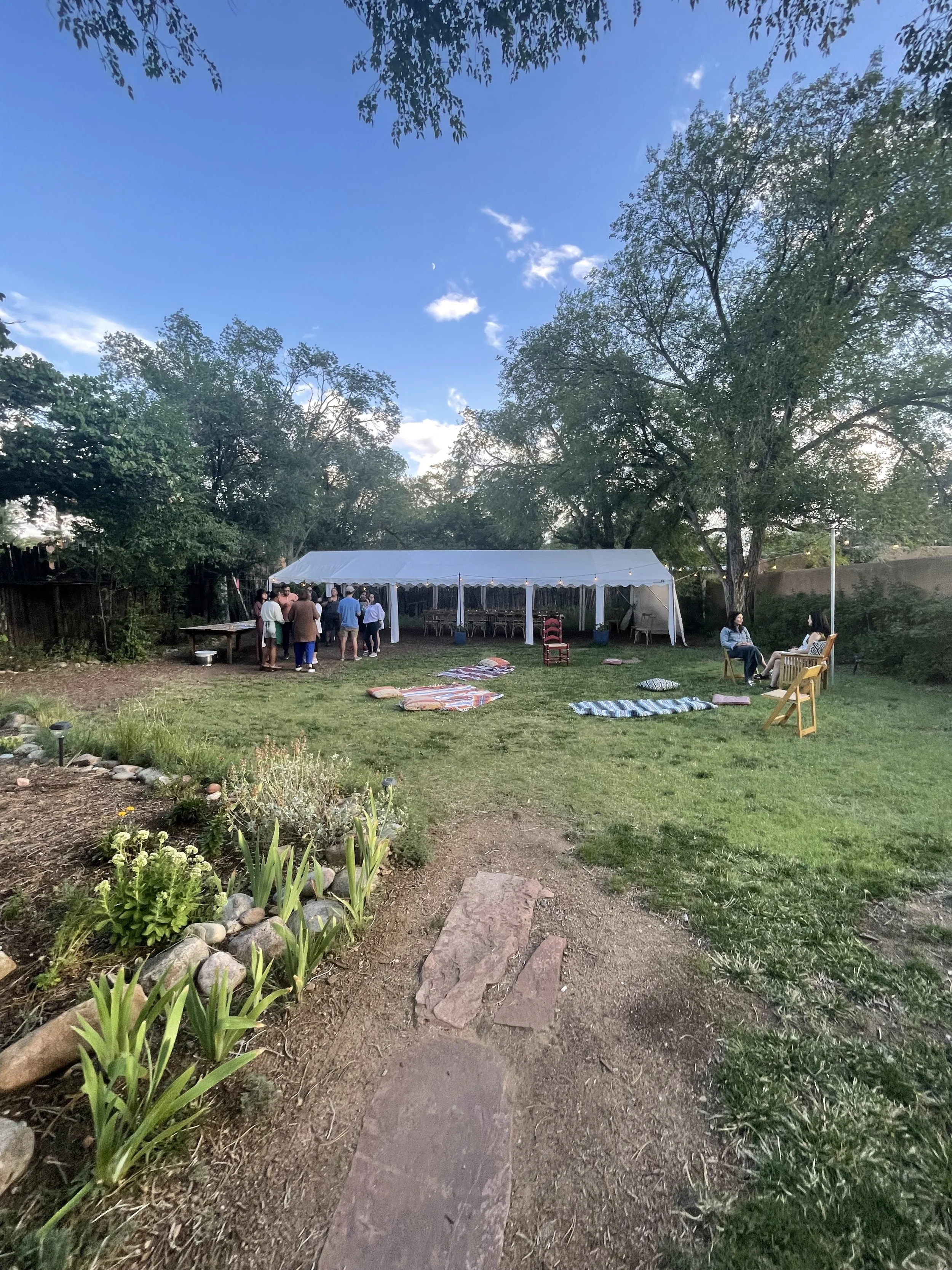 A backyard gathering under a white canopy tent with people socializing. There are chairs, blankets, and cushions on the grass, surrounded by trees and a clear blue sky.