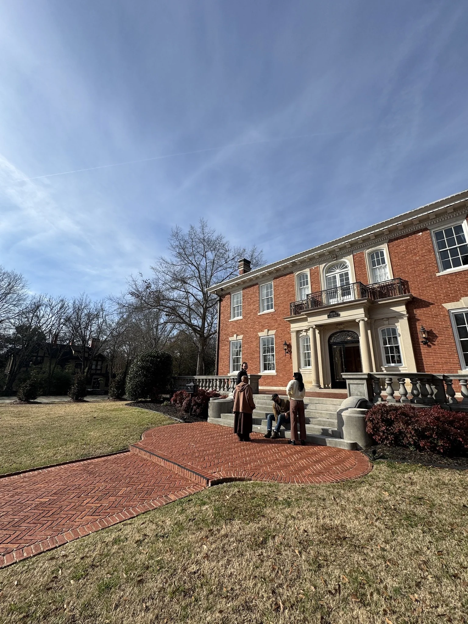 A group of people standing and sitting on the steps outside a large, historic brick mansion with a manicured lawn and trees around.