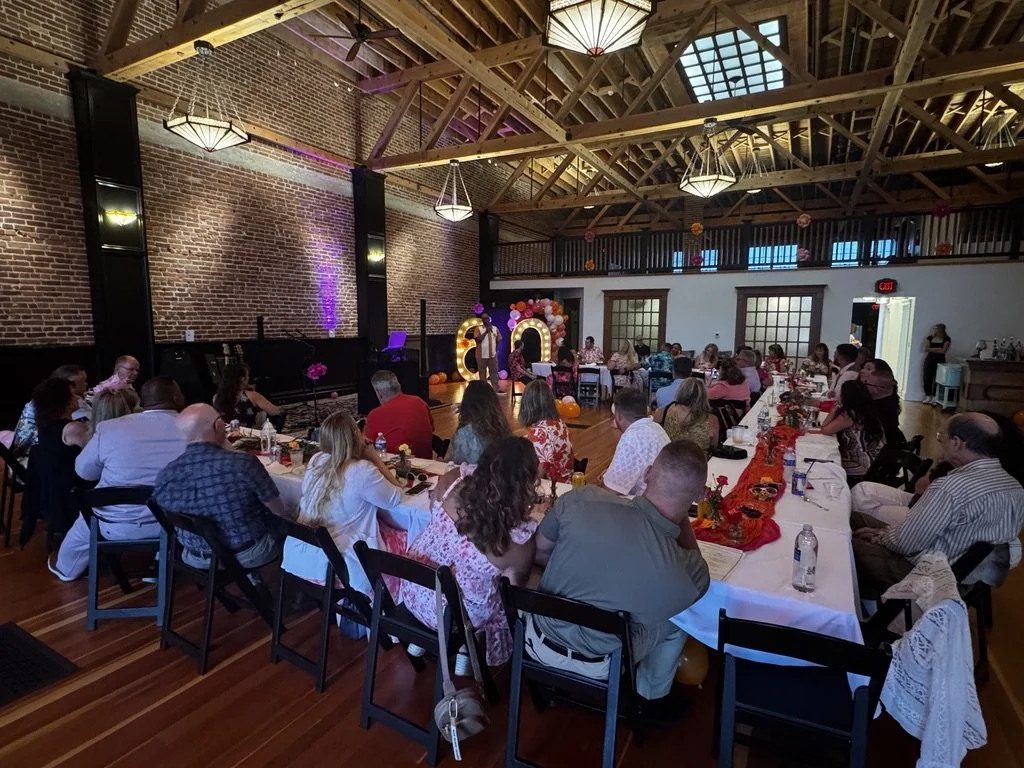 A large gathering of people seated at long decorated tables in a rustic event hall with exposed brick walls and wood ceiling beams, celebrating in front of a small stage with balloon and light decorations.