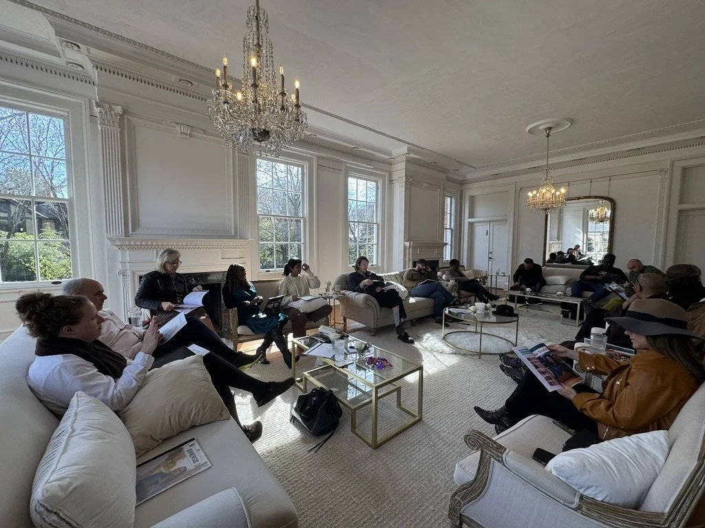 A group of people sitting on sofas and armchairs arranged in a semi-circle in a bright, elegant living room with large windows, chandeliers, and ornate white moldings, engaged in a meeting or discussion.