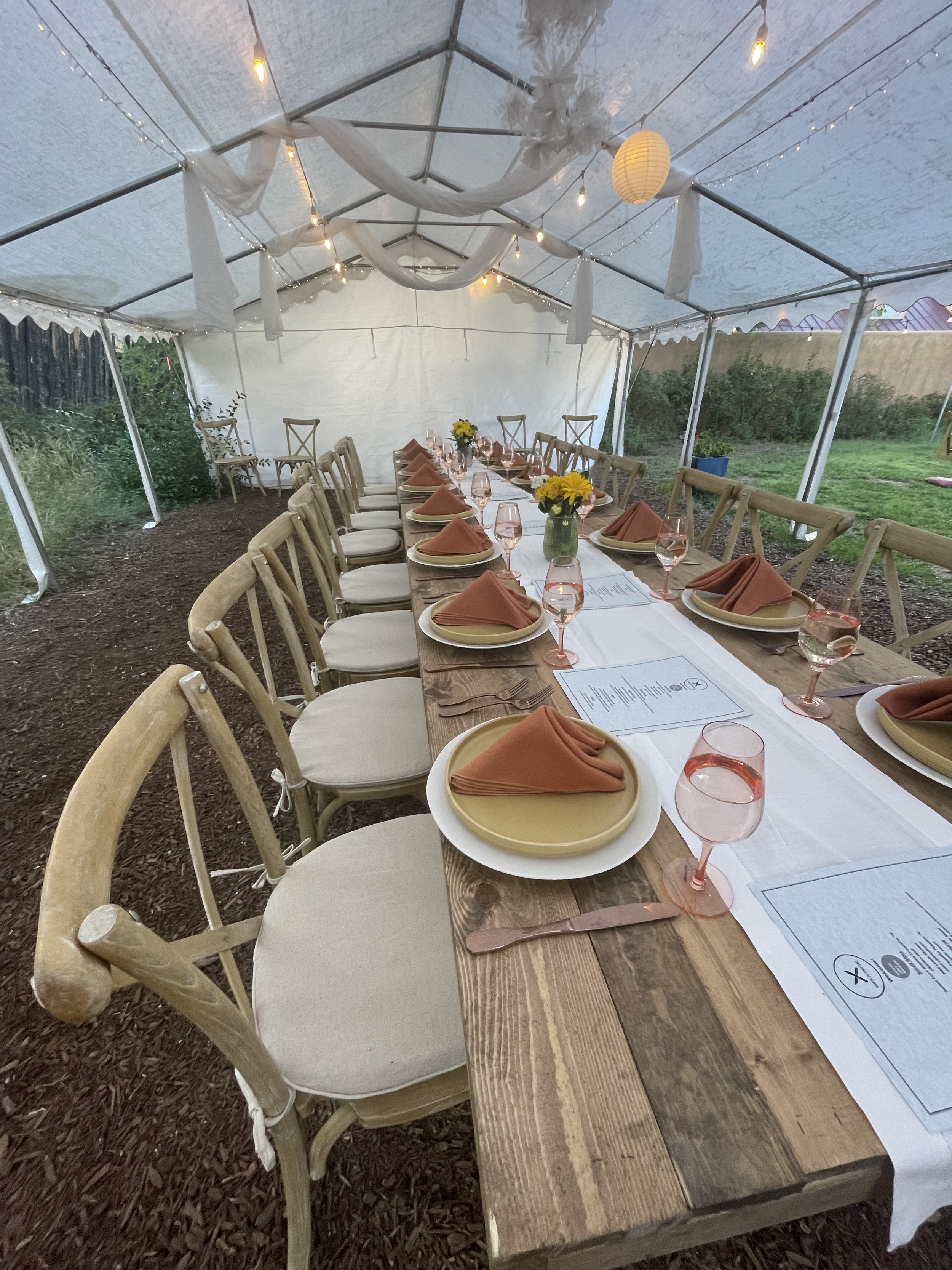 Table set for an outdoor party inside a white tent with string lights, decorated with yellow flowers in vases, with place settings including beige plates, rust-colored napkins, and pink wine glasses.