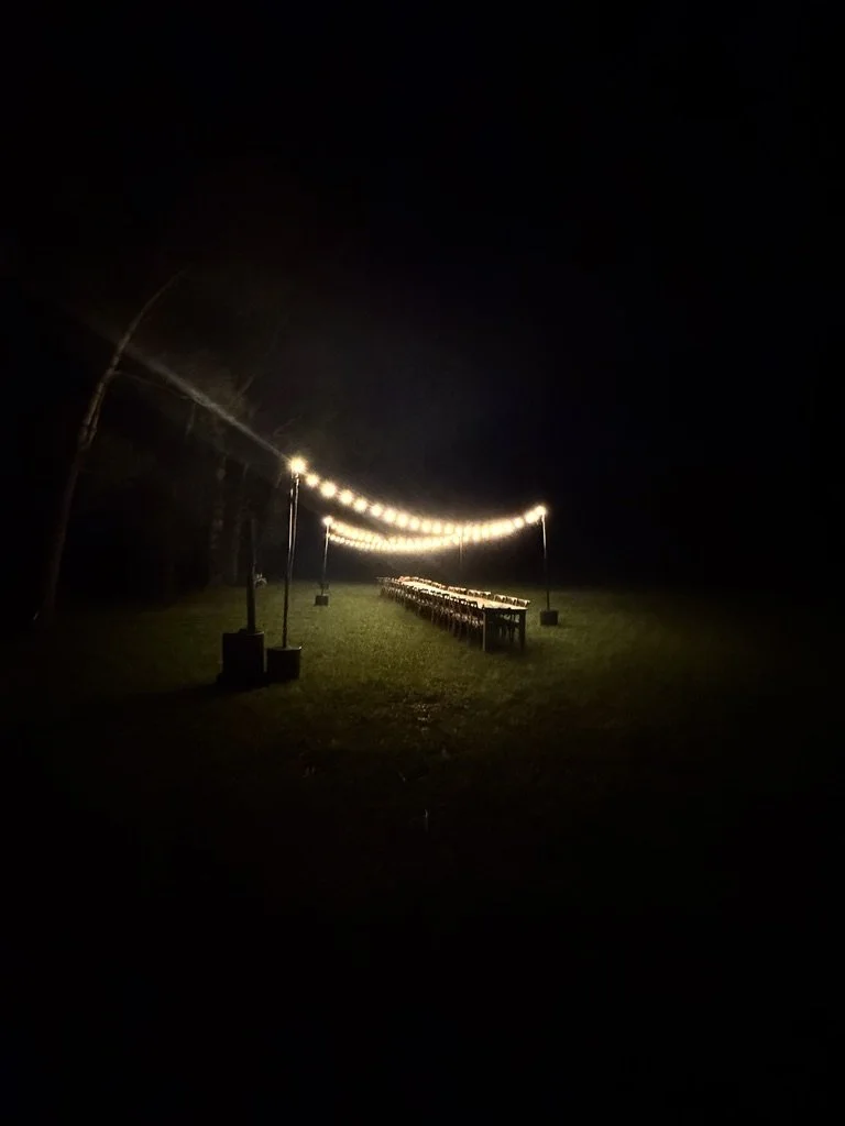 Nighttime outdoor scene with string lights hanging above a row of tables on grass.